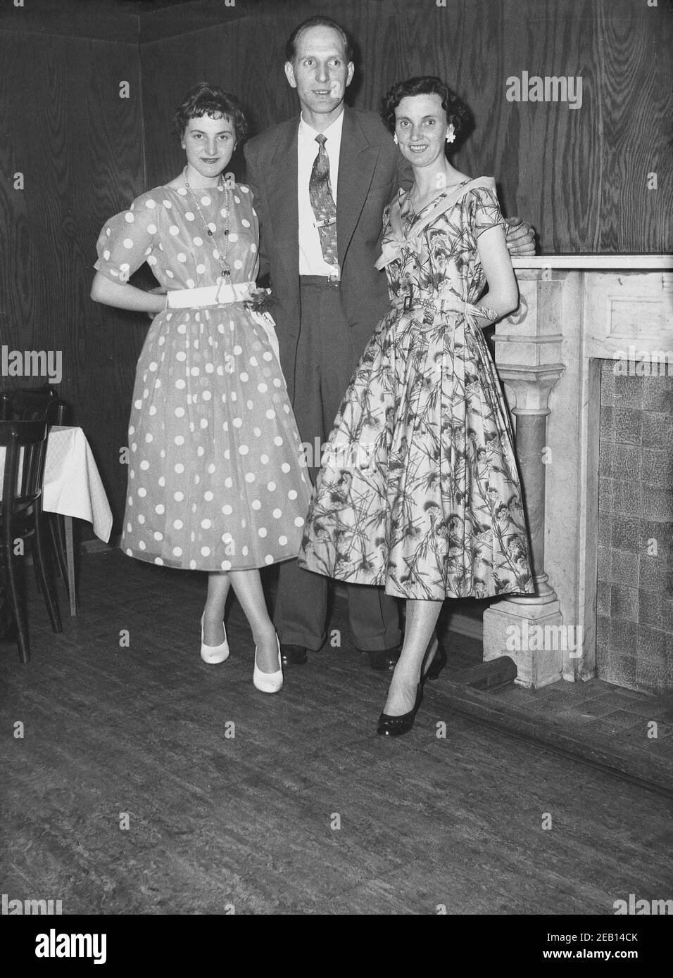 Años 50, histórica, joven dama en un vestido de topos o lunares de moda en esta época de pie para una foto con una pareja en su cumpleaños pary en una sala de eventos de hotel, Inglaterra, Reino Unido. Foto de stock