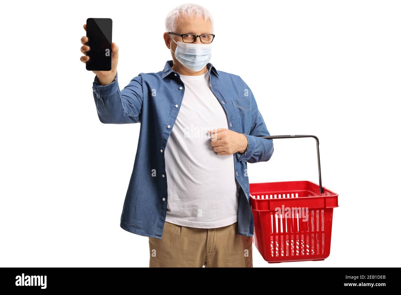 Hombre maduro con una cesta de la compra con un teléfono móvil y usando una máscara protectora aislada sobre fondo blanco Foto de stock