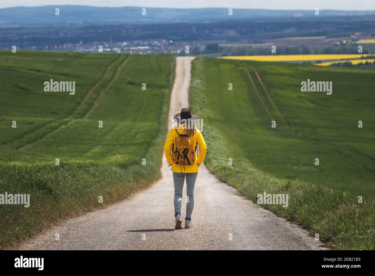 Largo viaje. Mujer de senderismo caminando en camino vacío. Hiker con sombrero y mochila con chaqueta impermeable amarilla. Concepto de viaje. Alejándose de ella Foto de stock