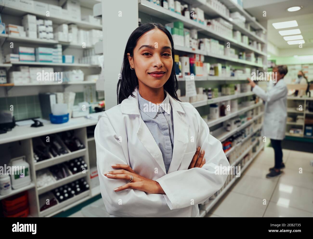 Mujer joven farmacéutica con las manos cruzadas y bata de laboratorio en el químico con un colega trabajando en segundo plano Foto de stock