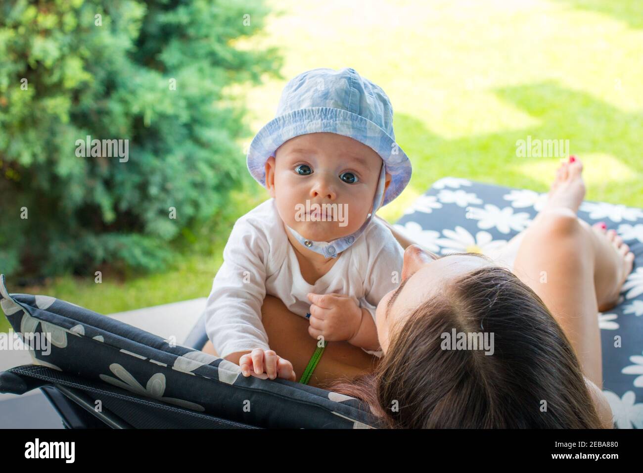 Madre joven con bebé en la gorra de sol tumbado en la silla de cubierta patrón de flores y jugando al aire libre juntos, verde natural patio trasero vista en el fondo Foto de stock