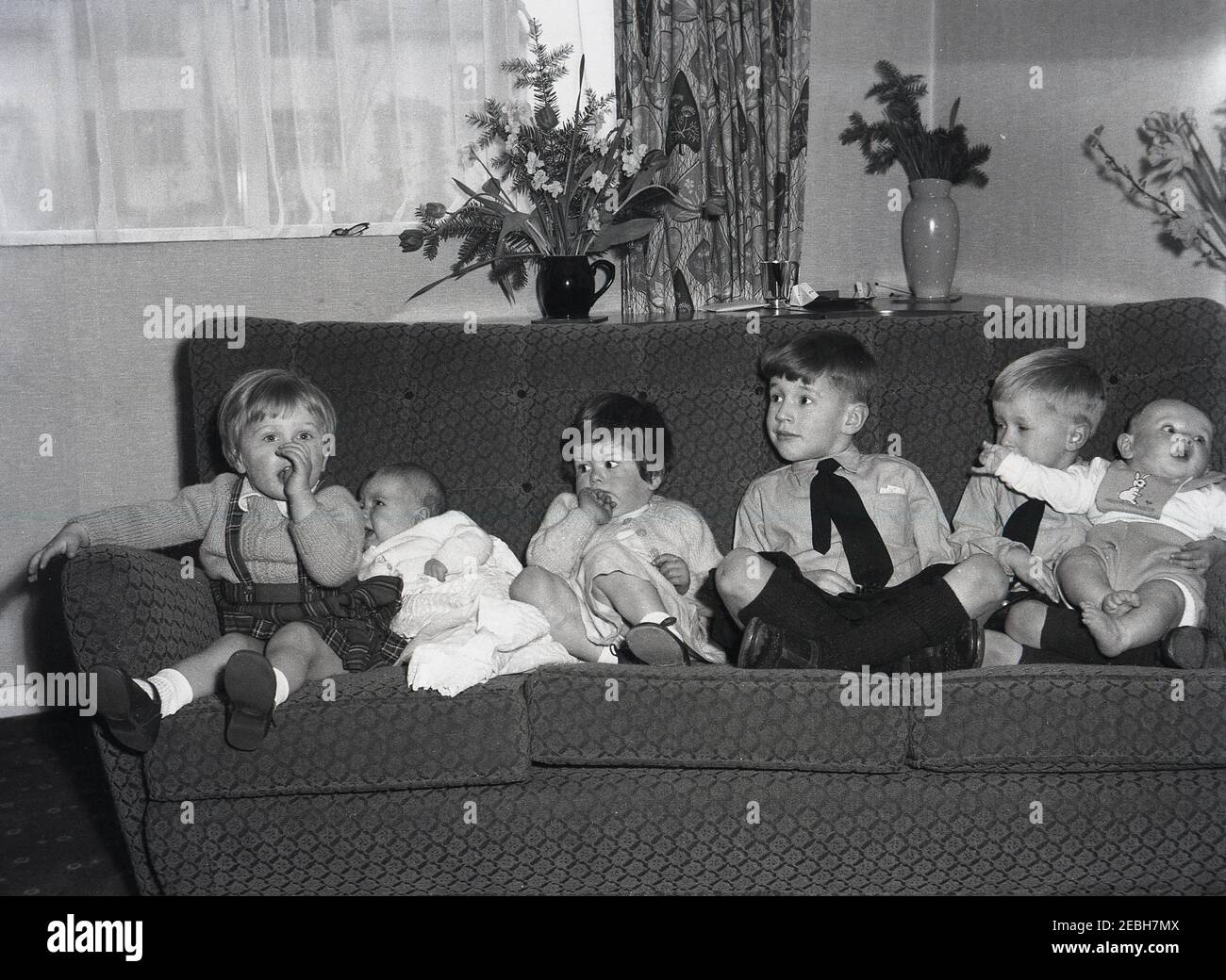 Años 50 histórico, en una habitación frontal, en un sofá, seis niños pequeños sentados juntos en una fila, dos niños pequeños, dos niñas y dos bebés, tal vez para una foto familiar, Inglaterra, Reino Unido. Conseguir que seis niños se sentaran juntos uno al lado del otro fue un logro. Foto de stock