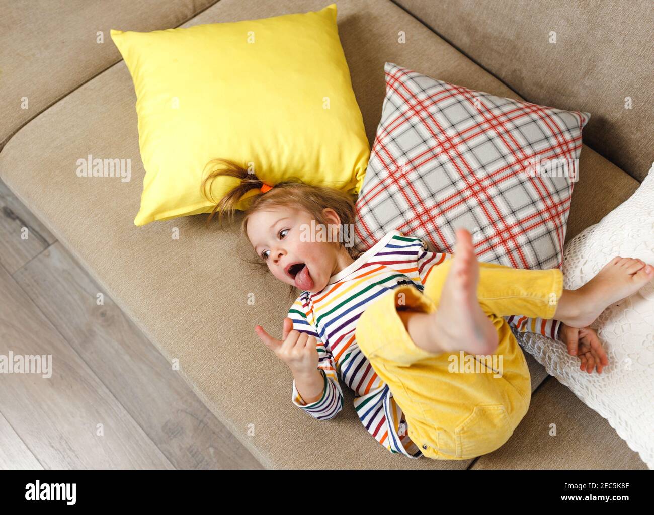 Una niña en pantalones amarillos y una camiseta de colores se encuentra en  su espalda en el sofá y muestra su lengua, haciendo diversión de ella.  Juegos para niños en el hogar.