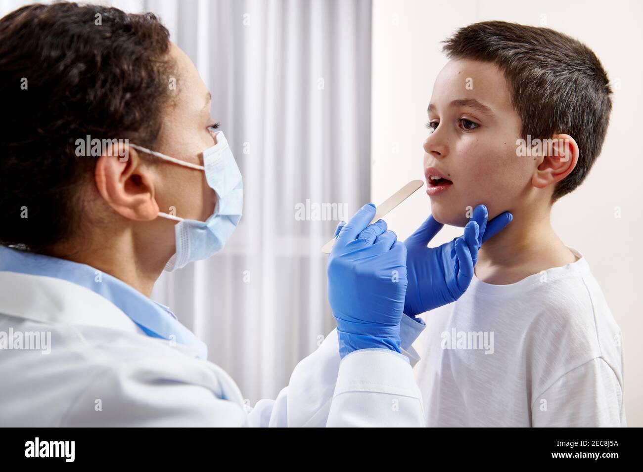 Médico afroamericano con máscara facial y guantes protectores que examinan la garganta del niño durante una visita domiciliaria. Foto de stock