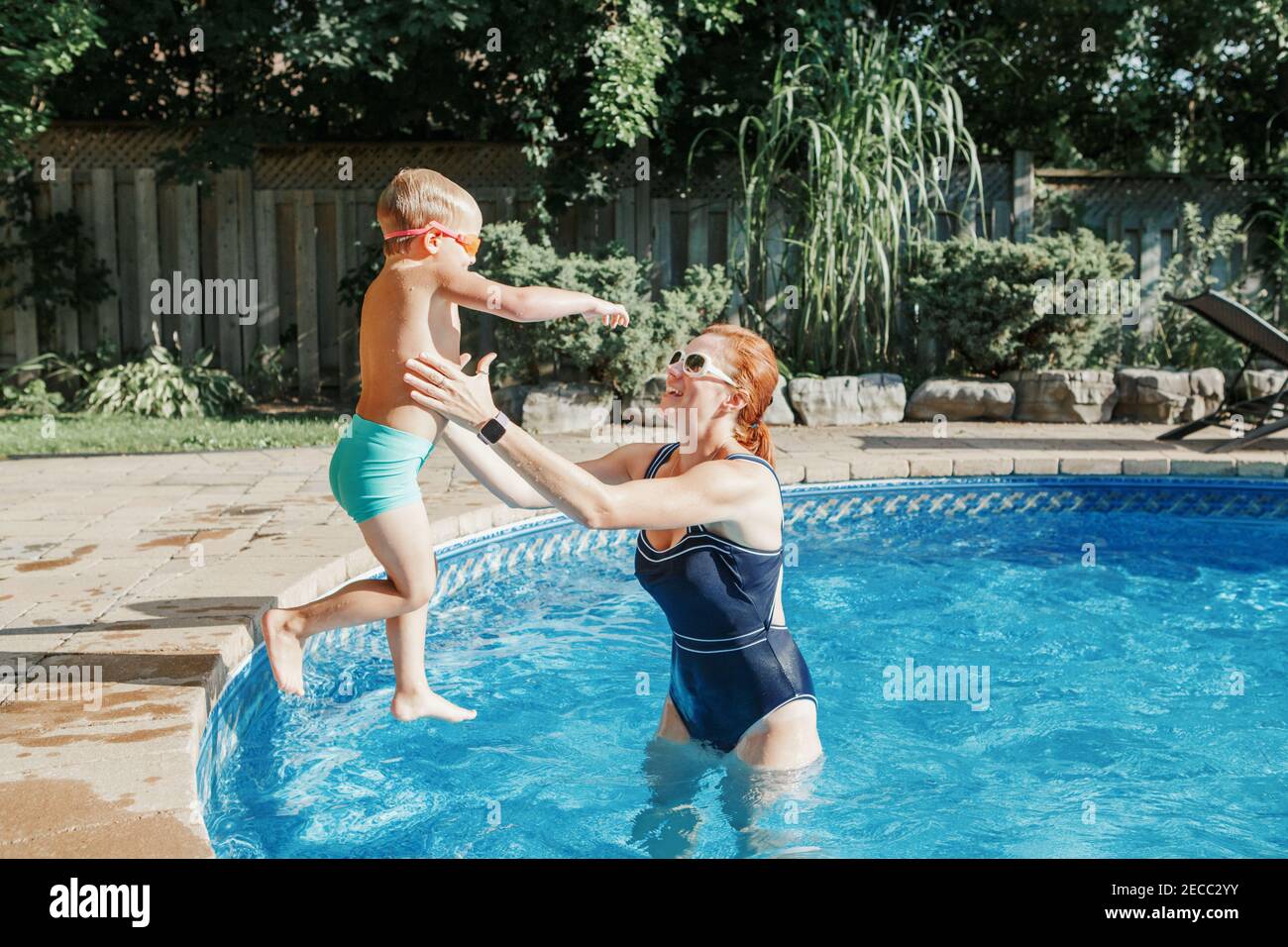 Madre caucásica entrenando a su hijo pequeño para nadar en la piscina al aire libre. Niño zambulléndose en el agua con gafas. Actividad estacional de la familia del agua Foto de stock