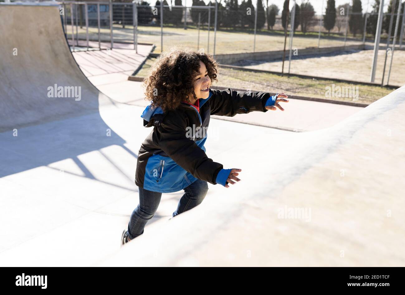 Vista lateral de un niño étnico positivo con peinado afro corriendo sube la rampa mientras te entretiene en el parque de patinaje durante el fin de semana día soleado Foto de stock
