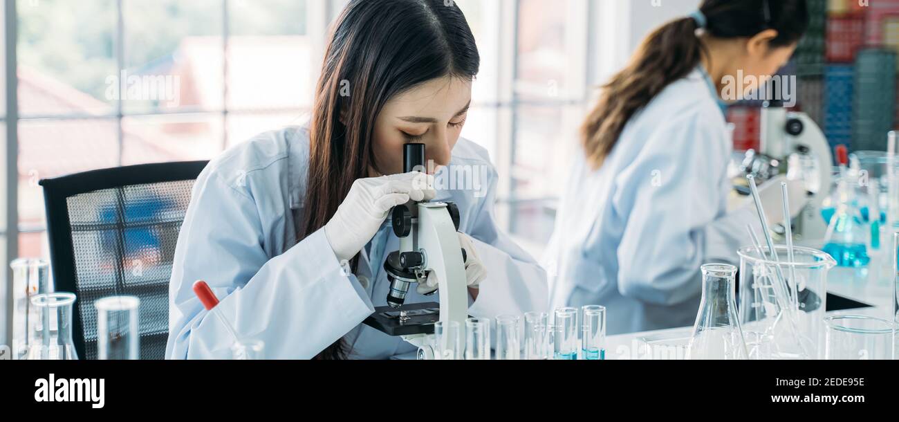 Joven científica asiática que hace investigación en laboratorio que usa laboratorio abrigo y guantes con colega en el fondo para desarrollar antivirus vacuna Foto de stock