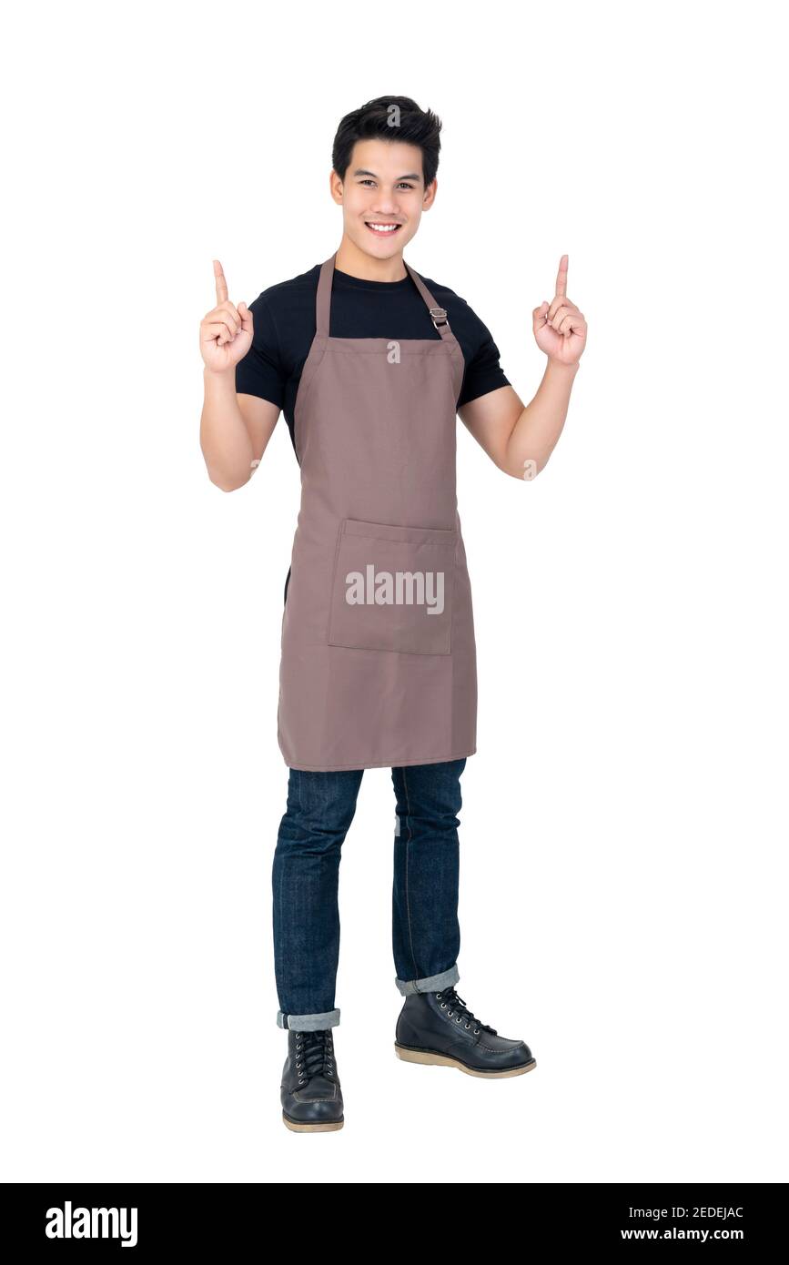 Retrato aislado de un joven y atractivo barista asiático en delantal marrón sonriendo y apuntando con la mano hacia arriba Foto de stock