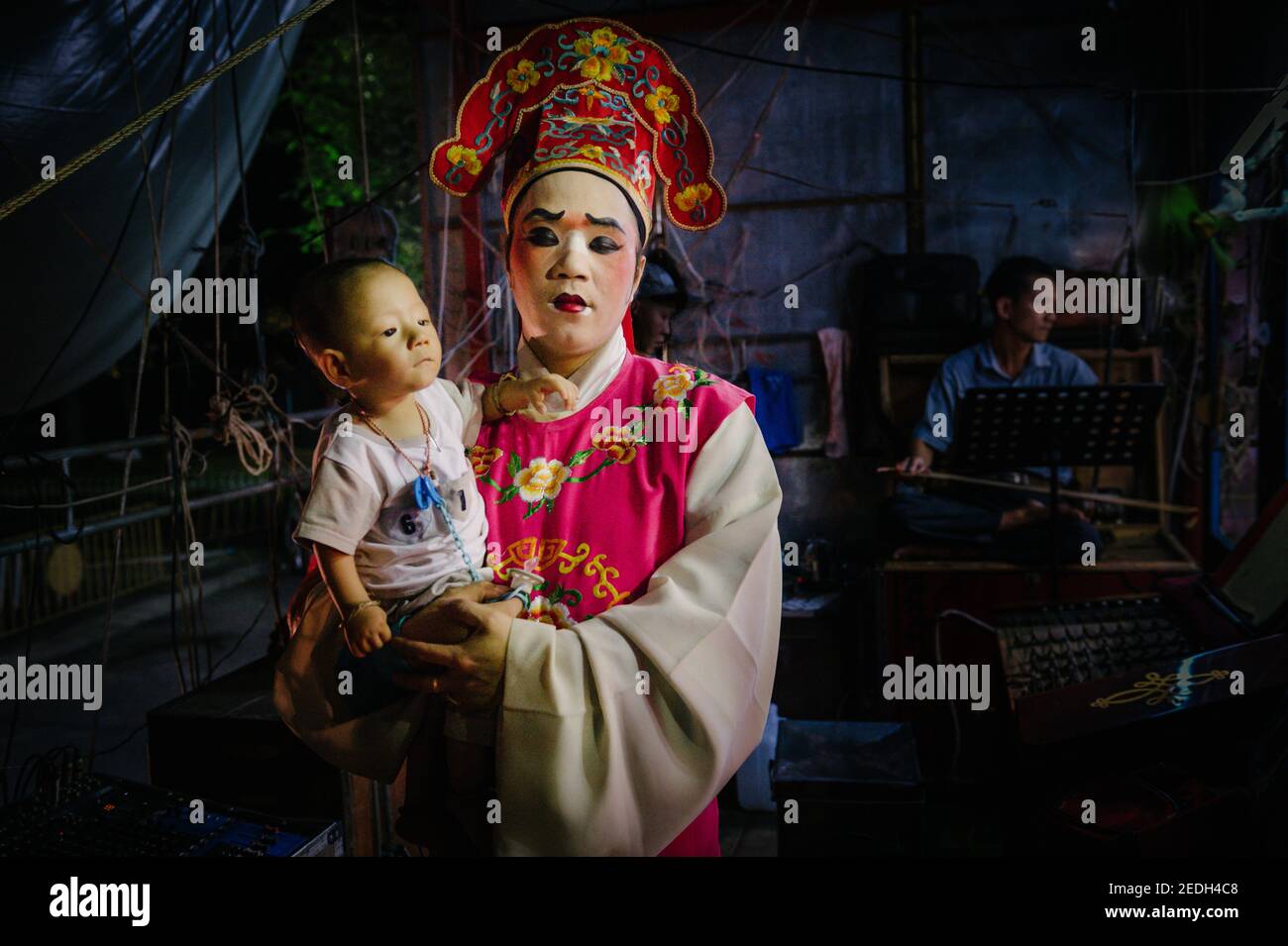 Un jugador de ópera chino completamente vestido con su colorido traje, sostiene a su bebé mientras espera entre bastidores para ponerse en el escenario. Foto de stock