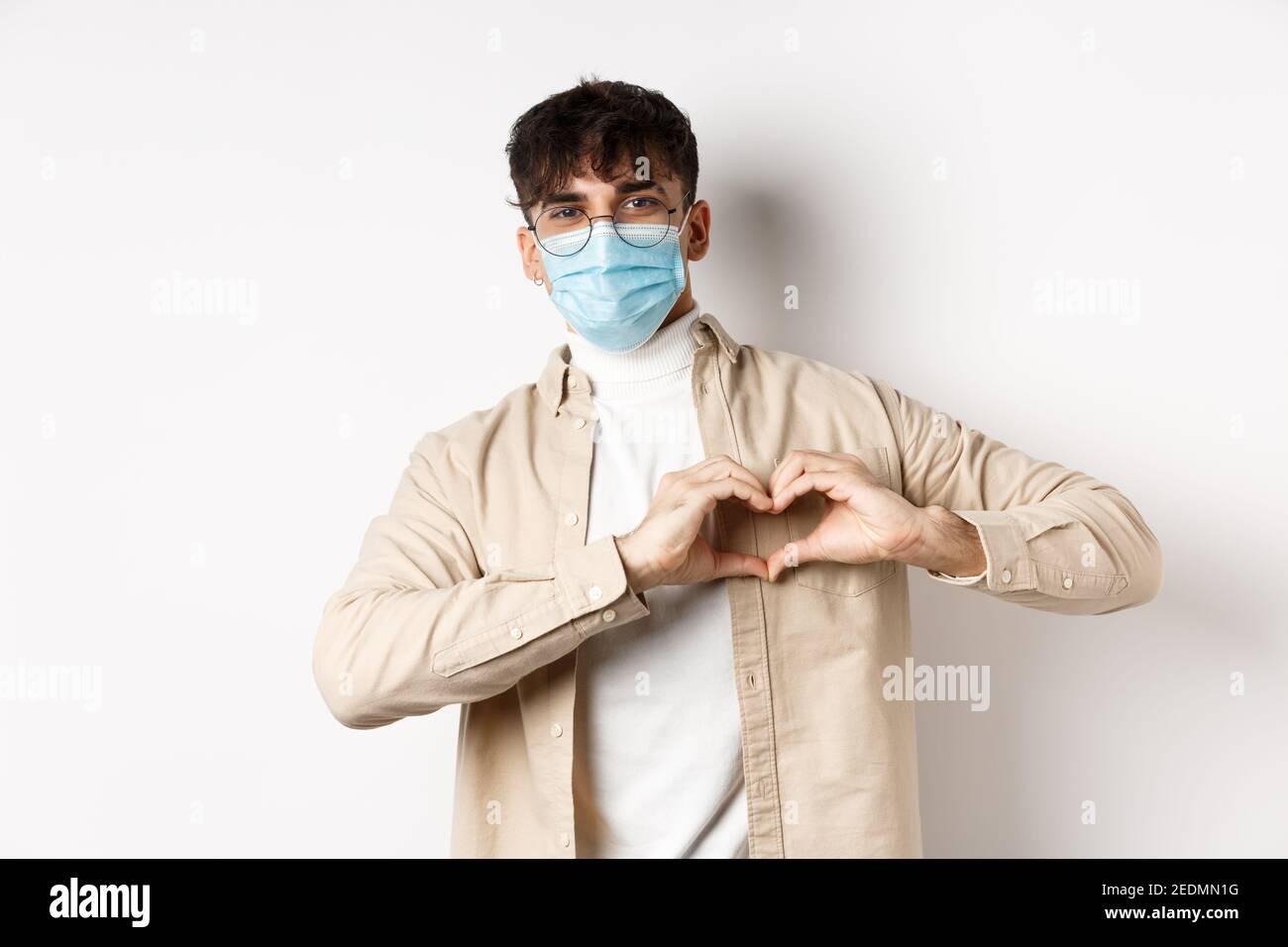 Concepto de salud, covid y cuarentena. Joven romántico en máscara médica estéril mostrando el gesto del corazón en el pecho, digamos que te amo, de pie en blanco Foto de stock