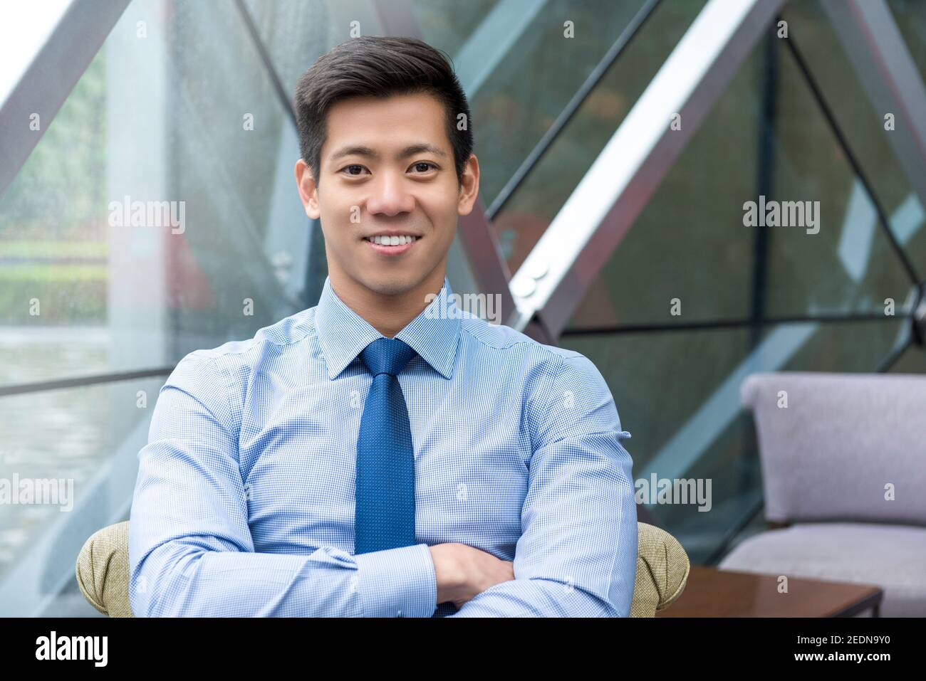 Retrato de un joven y sonriente hombre de negocios asiático tailandés-chino sentado con brazo cruzado en el salón de la oficina Foto de stock