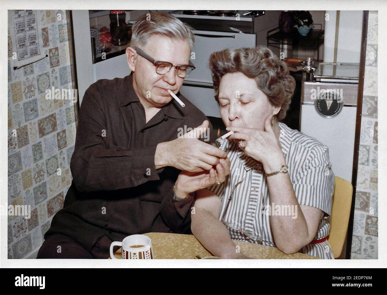 Una impresión fotográfica de la década de 1960 muestra a un hombre americano de mediana edad usando un encendedor de gas desechable para encender un cigarrillo para su esposa antes de que encienda su propio cigarrillo con punta de filtro después de que la pareja cenara en su pequeña cocina. Fumar en los Estados Unidos se hizo cada vez más popular en el siglo XIX, impulsado por la invención de la máquina de laminación de cigarrillos en 1881, y poco después de la introducción de los fósforos portátiles de "seguridad". El consumo anual aumentó de un promedio de 54 cigarrillos fumados por cada adulto en el país en 1900 a un máximo de 4,345 en 1963. Foto histórica. Foto de stock