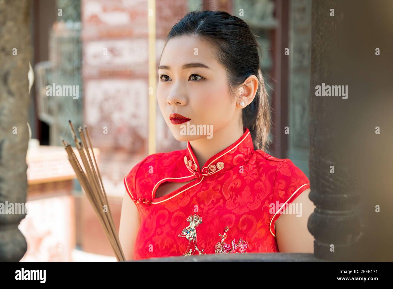 Mujer asiática en el tradicional vestido de qipao rojo rezando con incienso Palos durante el año Nuevo Chino o Lunar Foto de stock