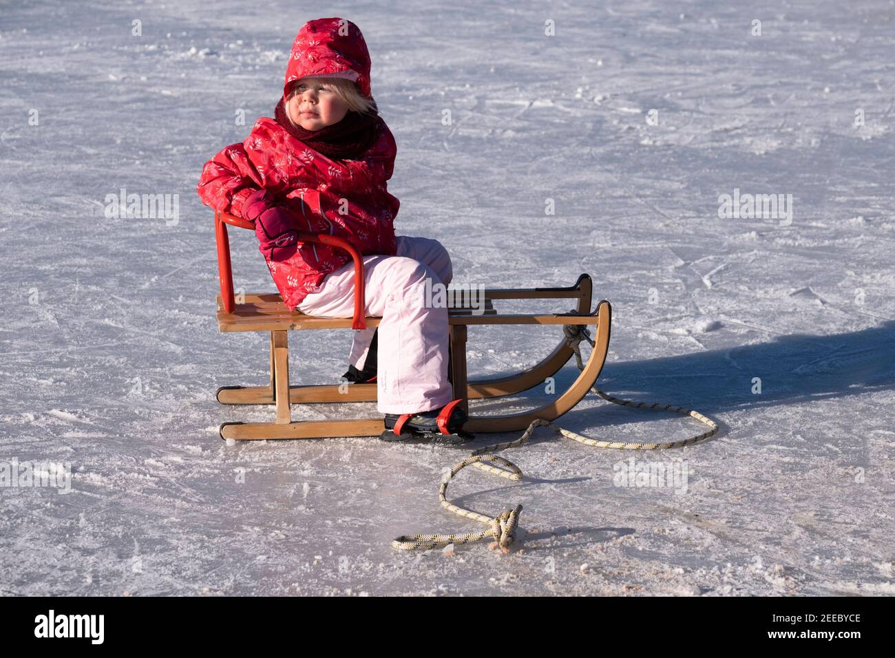 La niña está sentada en el camino en pantalones impermeables en un día frío  y soleado de invierno