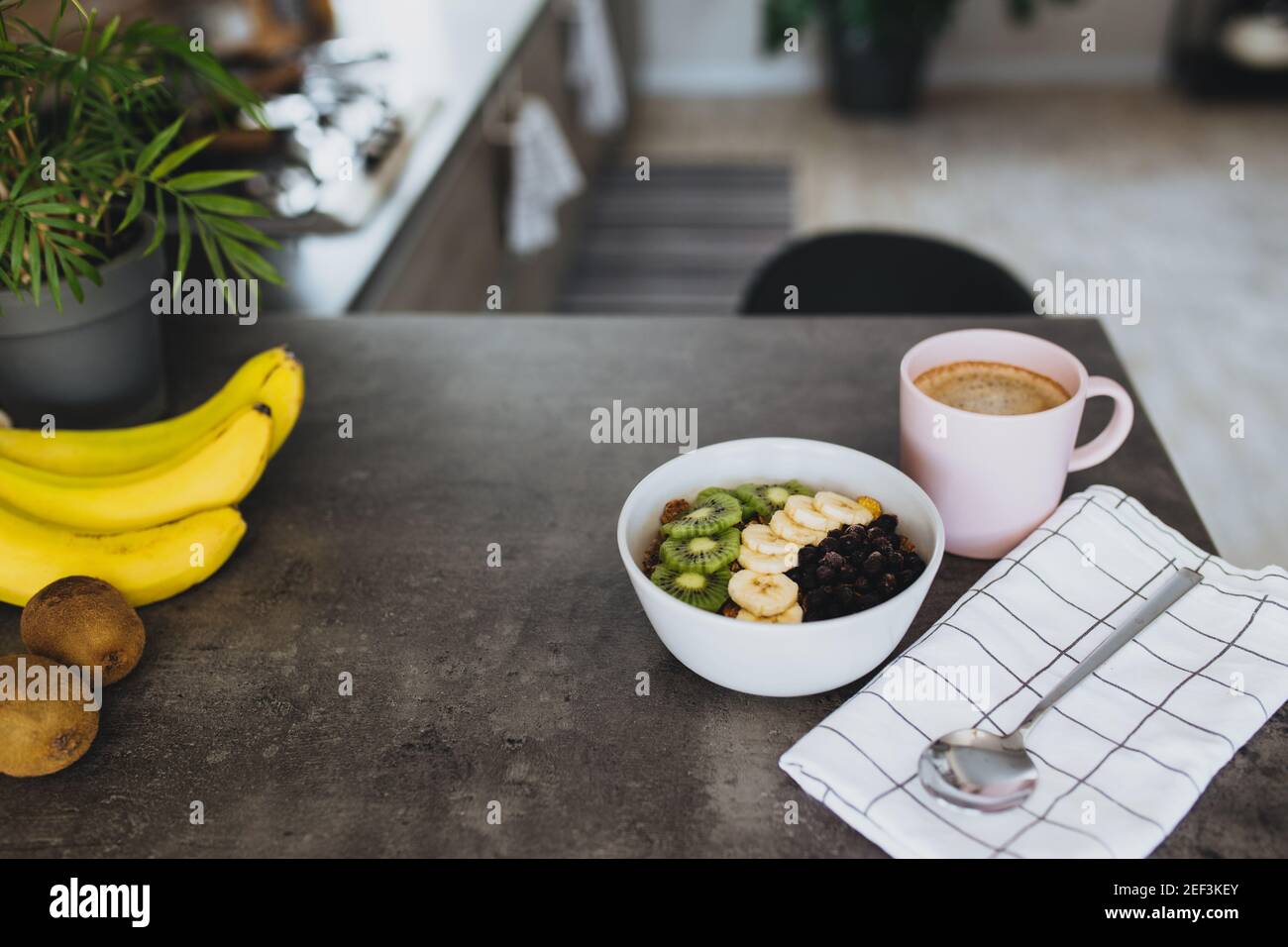 Taza de café rosa, cuenco con frutas tropicales picadas kiwi y plátano, arándanos, cuchara en barra en la elegante cocina tipo loft. Fondo borroso. Hig Foto de stock