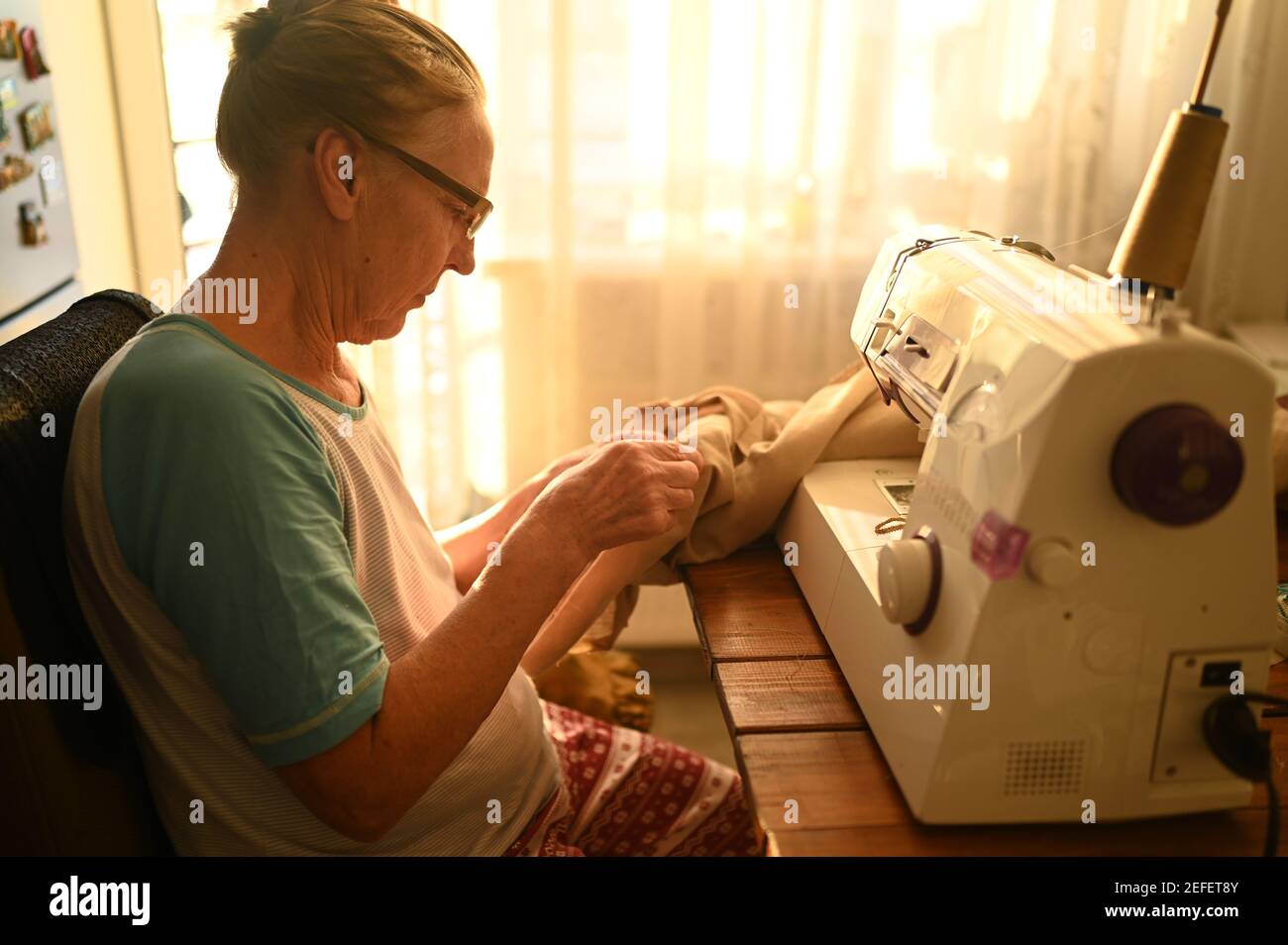Mujer anciana segura mujer costurera blanca camisa y gafas sentadas frente  a la máquina de coser, trabajando en la ropa en casa usando tela beige.  Jubilados, edad, ocupación de trabajo concepto Fotografía
