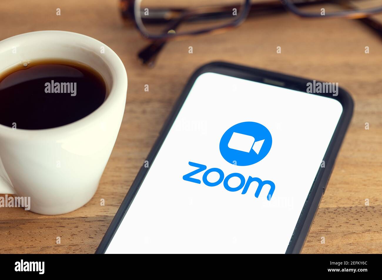 Galicia, España; 15 de febrero de 2021: Zoom logo en pantalla de teléfono inteligente sobre escritorio gafas y taza de café sobre mesa de madera Fotografía stock - Alamy