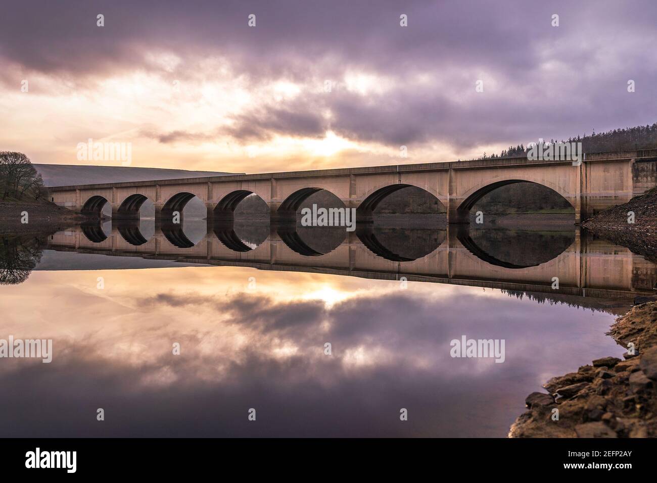 Largo puente arqueado cruzando el embalse de Ladybower reflejado en el Valle de Derwent todavía aguas profundas del río con el hermoso amanecer en el cielo naranja detrás Foto de stock