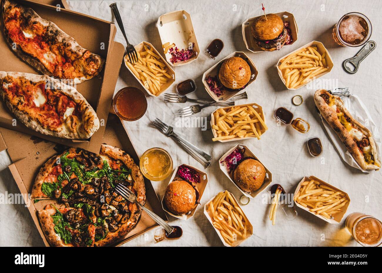 Cena de comida rápida cerrada desde el concepto de servicio de entrega. Piso-Lay de la fiesta de la casa de cuarentena con hamburguesas, patatas fritas, sándwiches, pizza, cerveza en vasos sobre blanco mantel fondo, vista superior Foto de stock