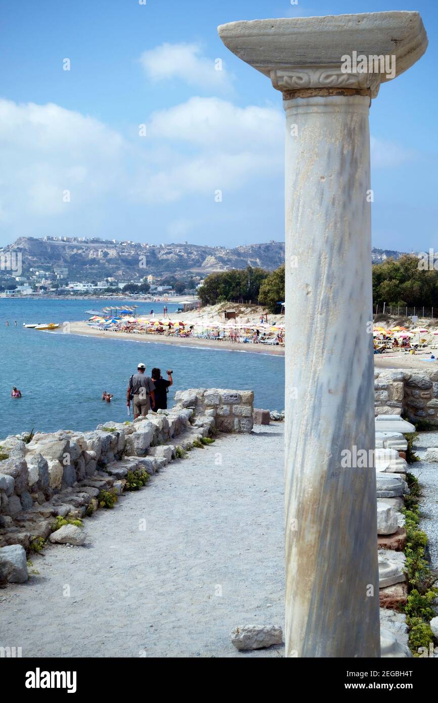 Un pilar de mármol de las ruinas de Agios Stefanos contrastan con los turistas, sombrillas y bañistas en la playa de Kefalos, Kos Foto de stock