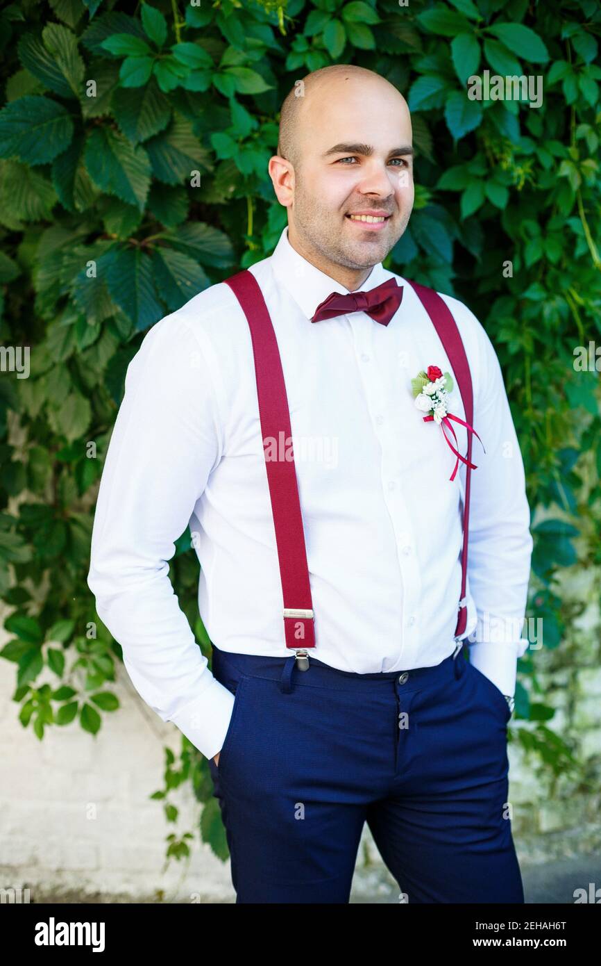 Retrato de un novio de hombre con un lazo rojo y tirantes. Él está seguro con una en su rostro. Elegante y moderno hombre barbudo en una camisa blanca Fotografía de