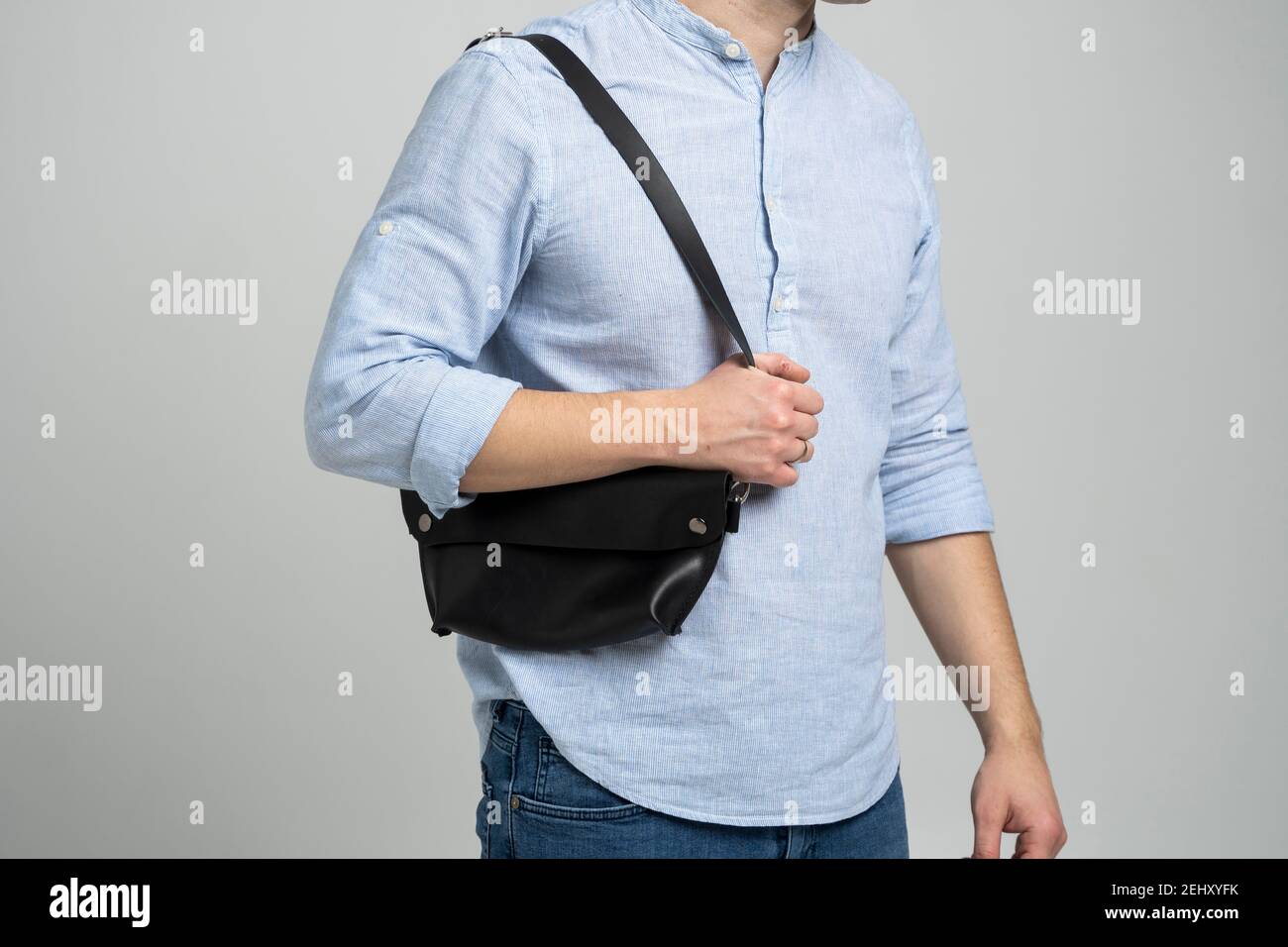Hombre en una camisa azul con una bolsa de cuero negro hecha a mano sobre  su hombro. Bolso de bananas oscuro. Hombre en un estudio. Cómoda bolsa  pequeña para caminar Fotografía de