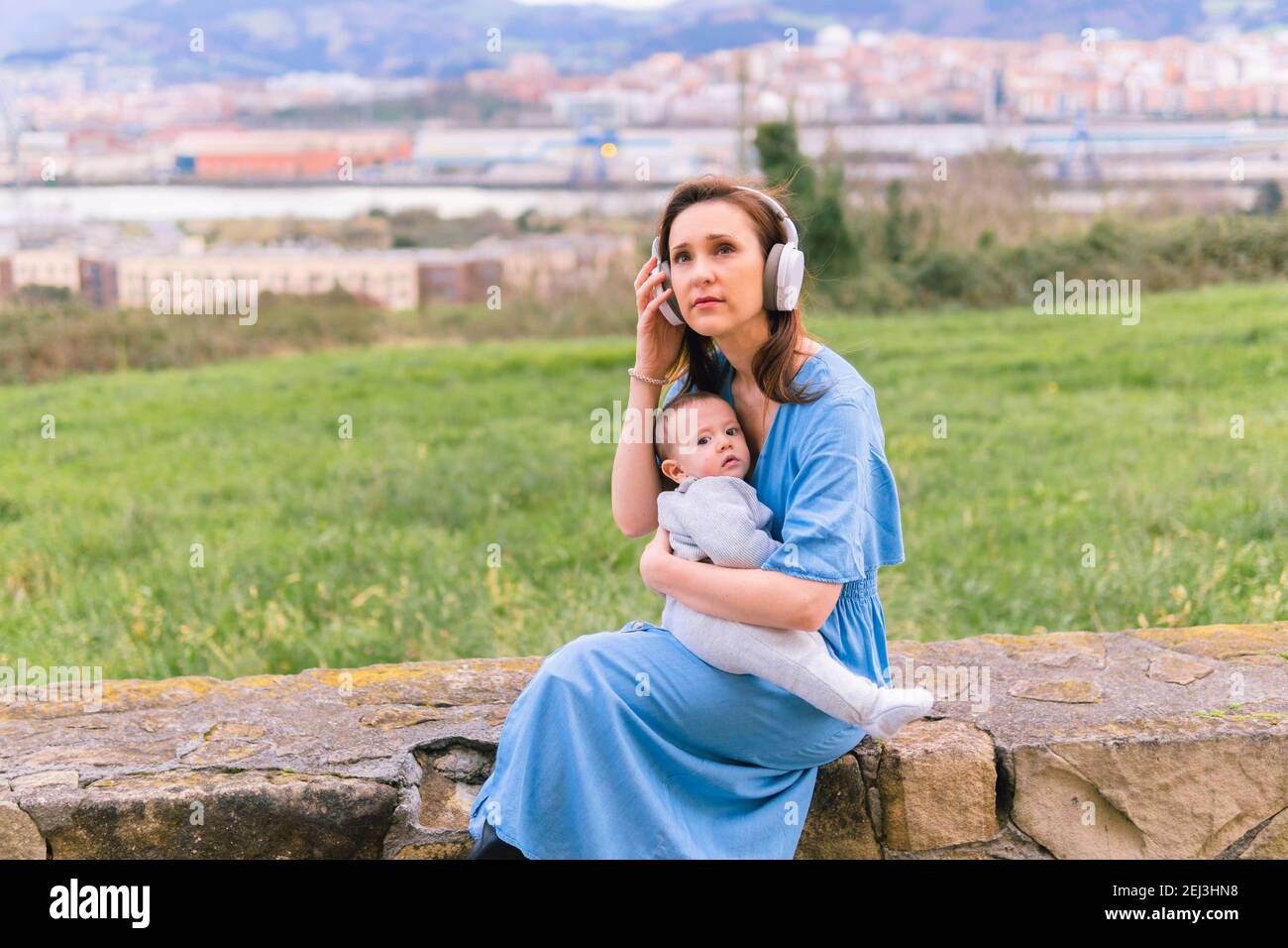 hermosa mujer joven de mediana edad con auriculares de música y vestido azul  besando a su bebé de seis meses en primavera en el campo. concepto de moda  Fotografía de stock -