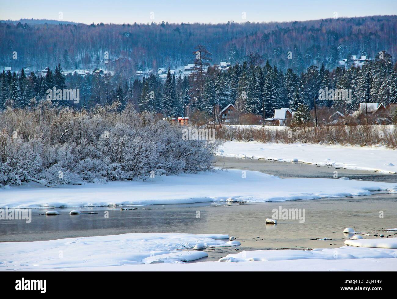 Varias cabañas con tejados cubiertos de nieve entre los árboles de coníferas del bosque. En el primer plano - un pequeño río hace un giro, Foto de stock