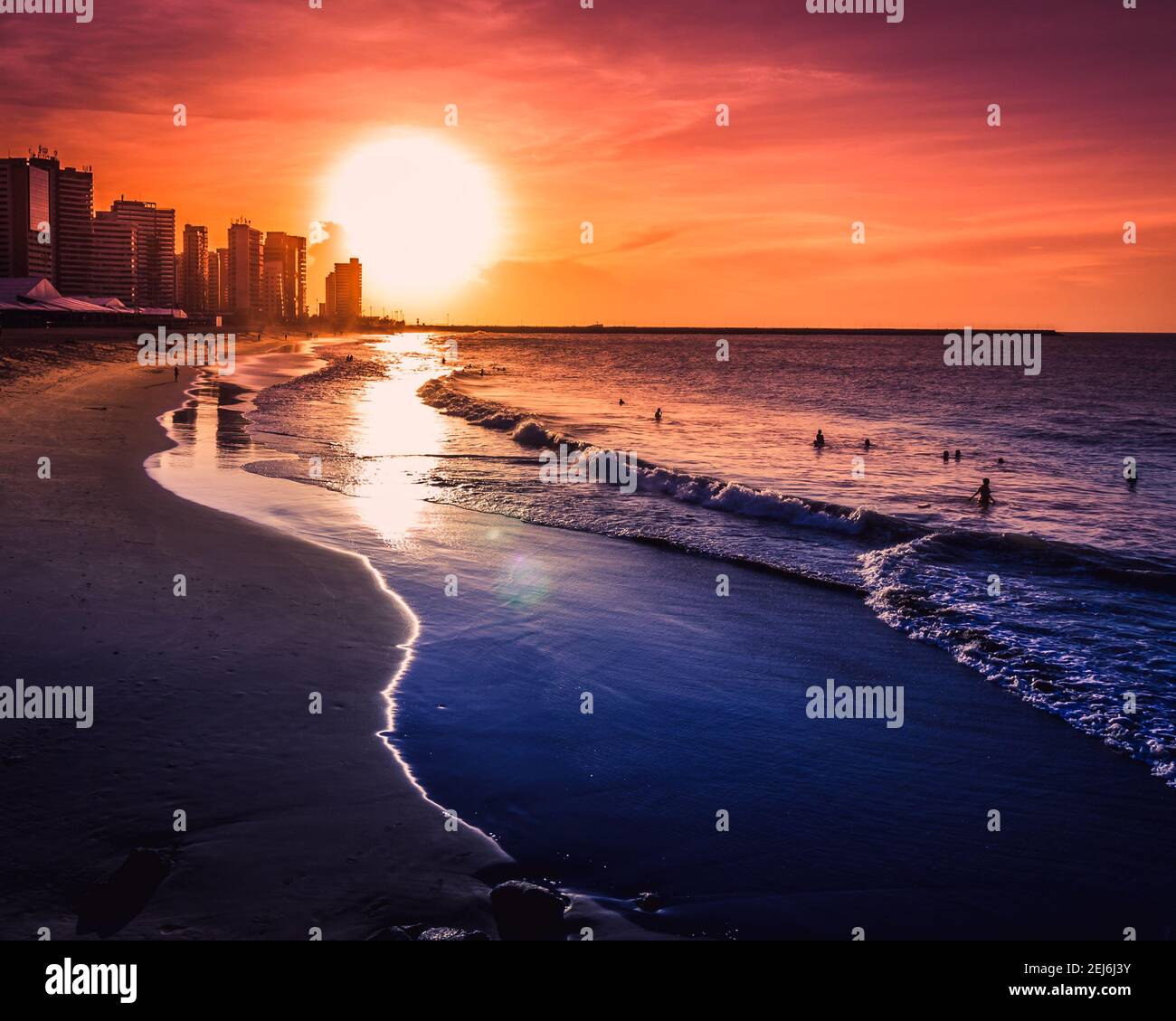 Una ardiente puesta de sol naranja en una playa tropical con algunos edificios y amantes de la playa bañándose en el mar cálido. Foto de stock