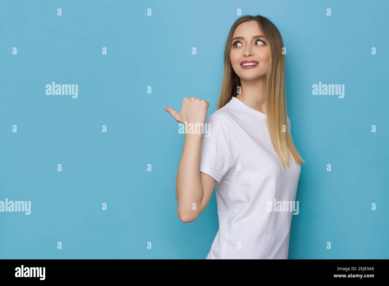 Sonriente mujer joven en camisa blanca está apuntando detrás de sí misma con el pulgar y mirando lejos. Vista lateral. Estudio de cintura arriba sobre fondo azul. Foto de stock