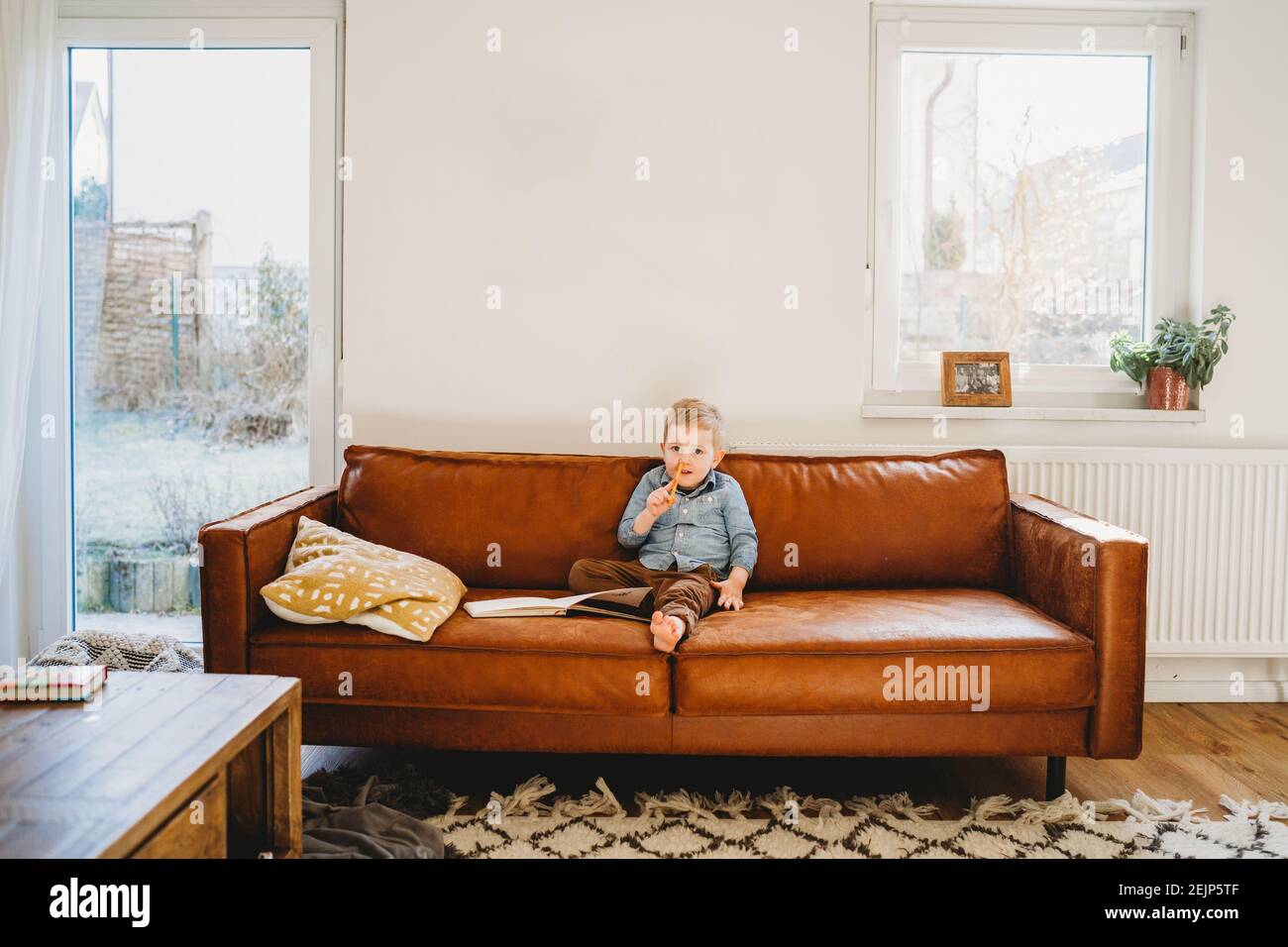 Adorable niño rubio blanco sentado en el sofá con lápiz su nariz Foto de stock