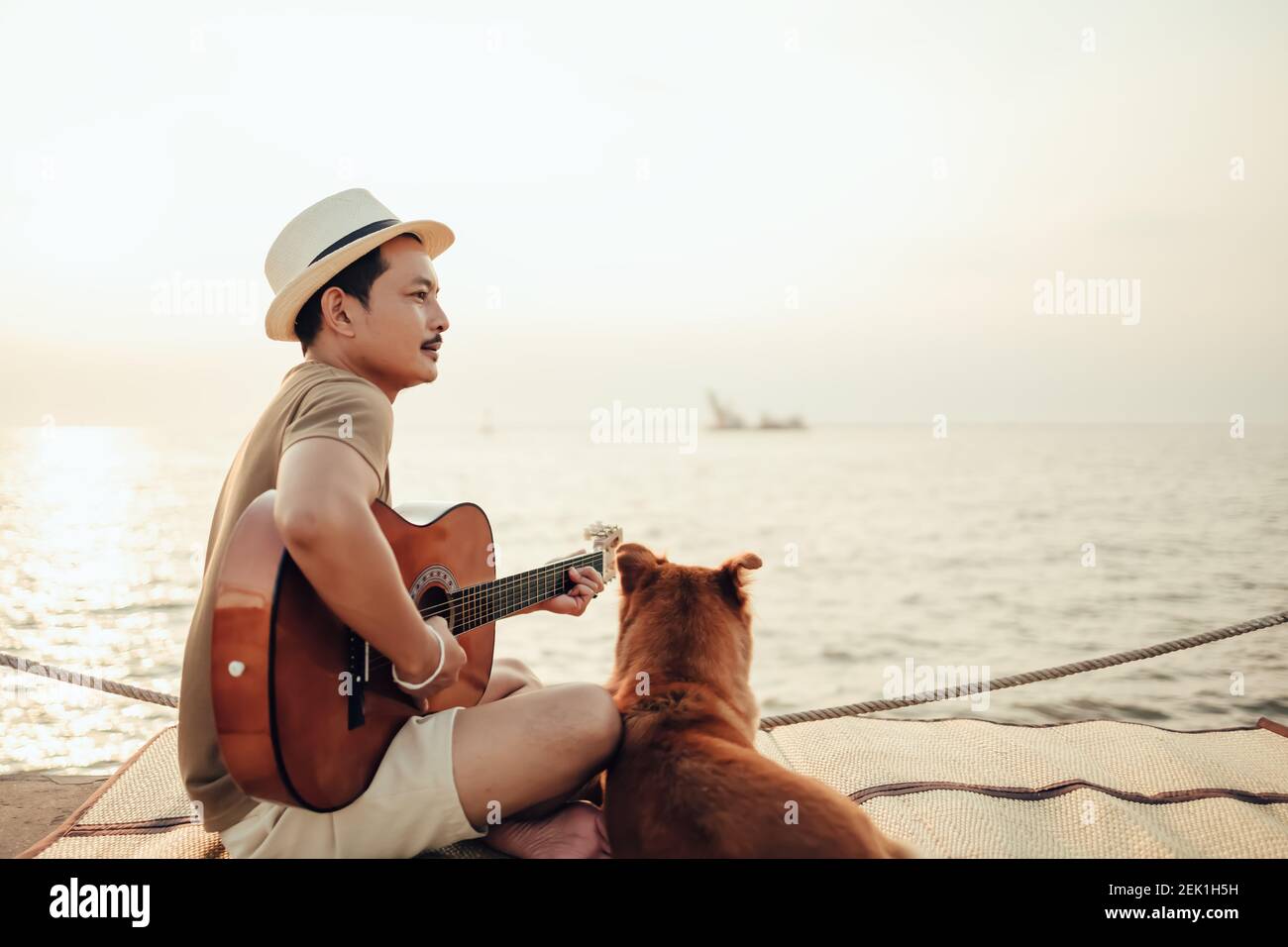 Rubí Microordenador a pesar de Un hombre lleva sombrero de paja y tocando música de guitarra cerca de la  puesta de sol del mar con una mascota de perro. Viaje, vocación, concepto  de vacaciones Fotografía de stock -