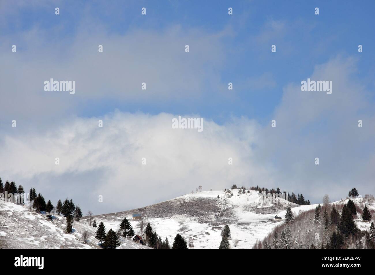 Paisaje escénico de colinas con pequeñas casas y árboles, cubierto de nieve y azul cielo nublado fondo Foto de stock