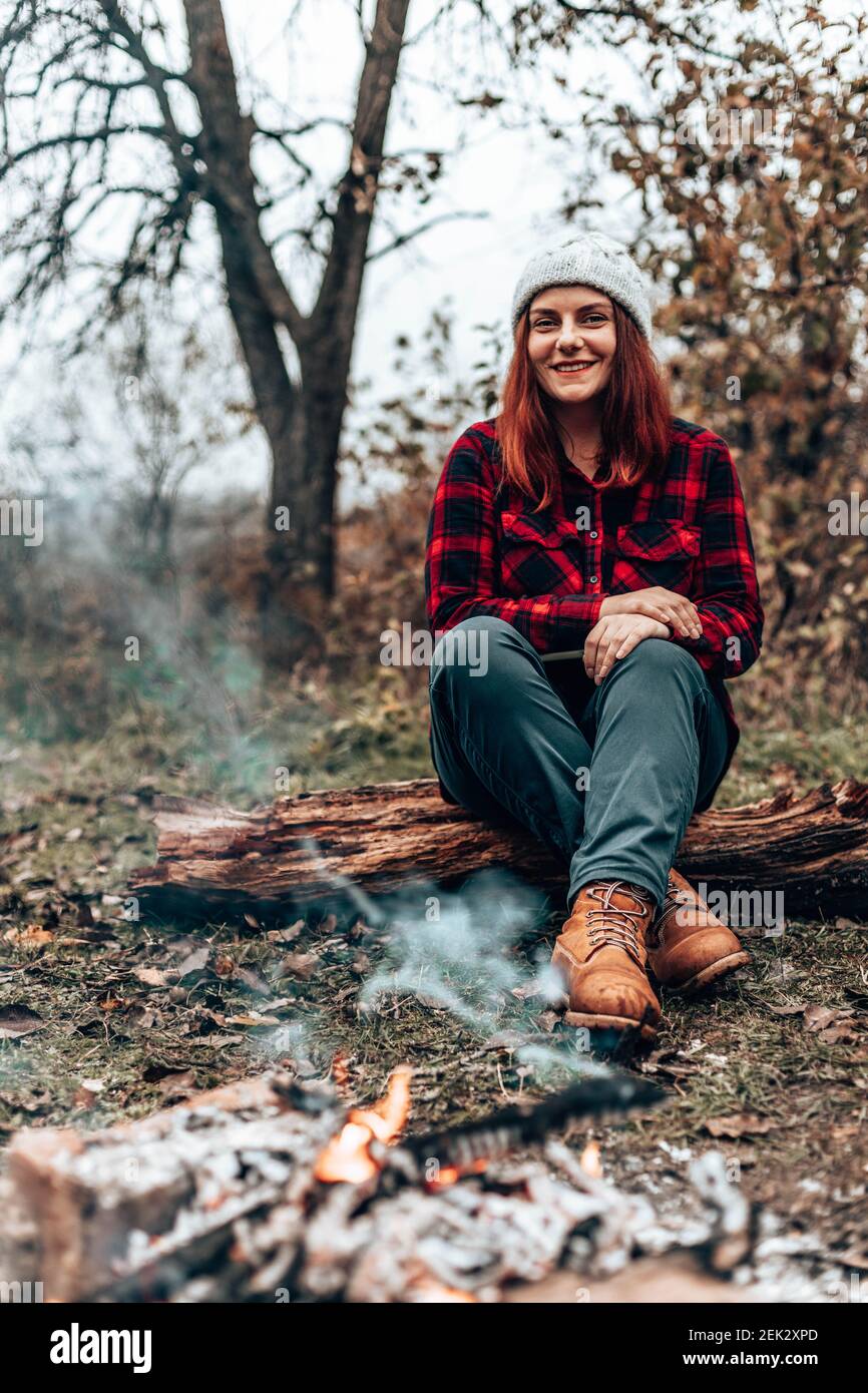 Feliz viajero niña con ropa de hipster se sienta en un tronco en el bosque cerca del fuego por camping. Mujer disfrutando relajándose en el bosque Foto de stock
