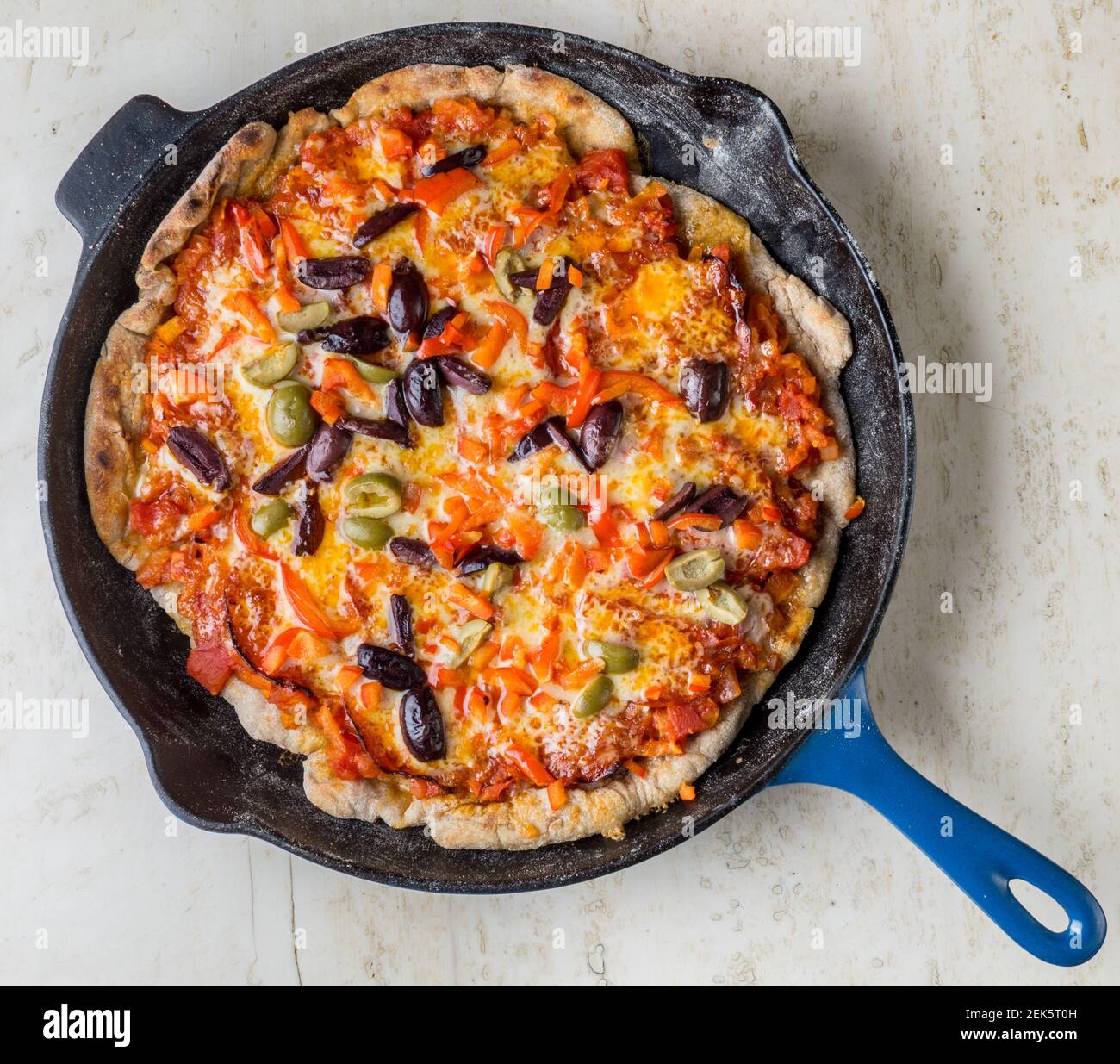 Una pizza casera a la parrilla con pasta de tomate, chorizo, oliva, queso cheddar y capsicum que se repone en una sartén azul le Creuset sobre una encimera de mármol Foto de stock