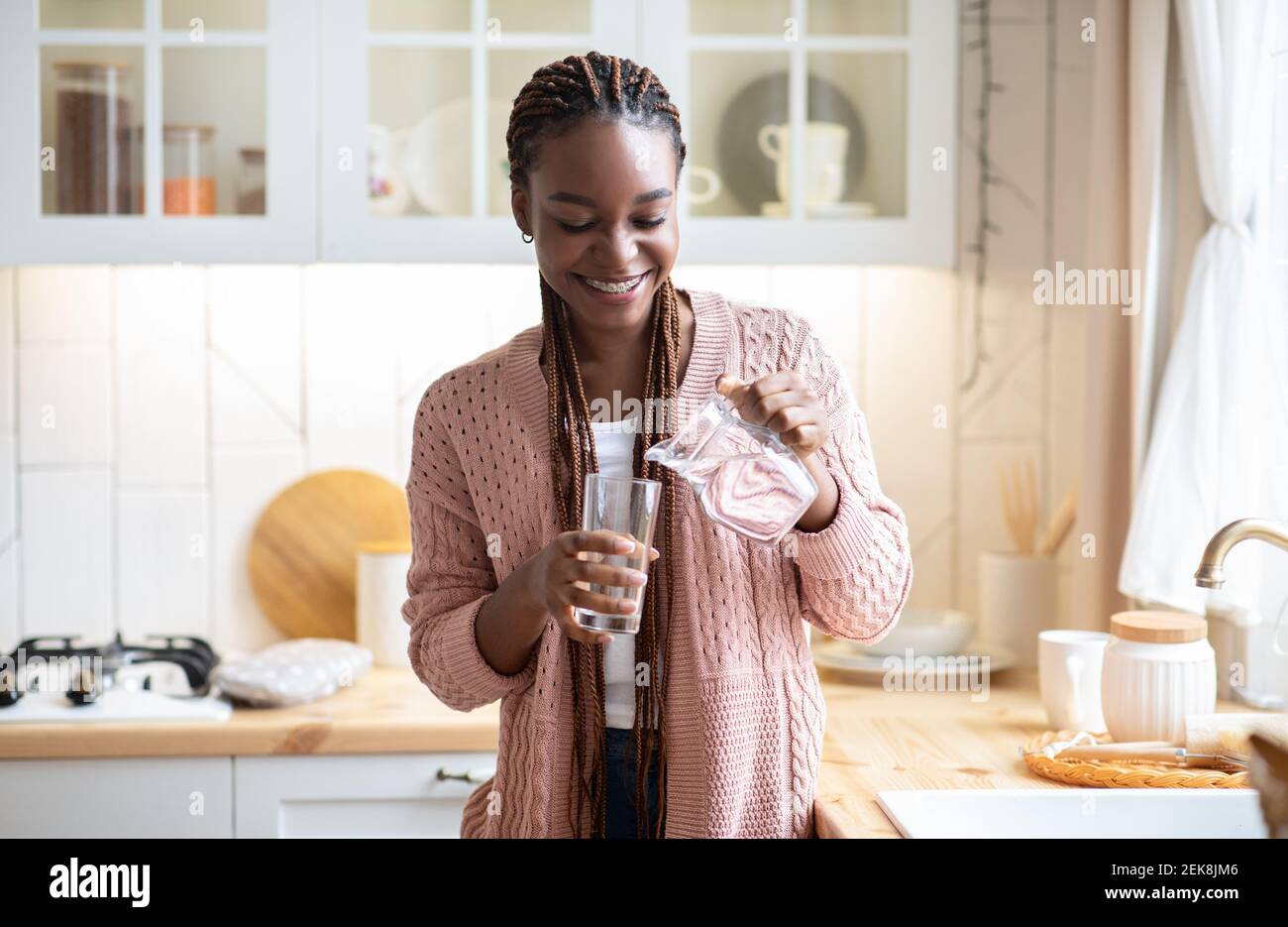 Mujer negra sedienta agua potable en la cocina, vertiendo líquido sano a vidrio Foto de stock