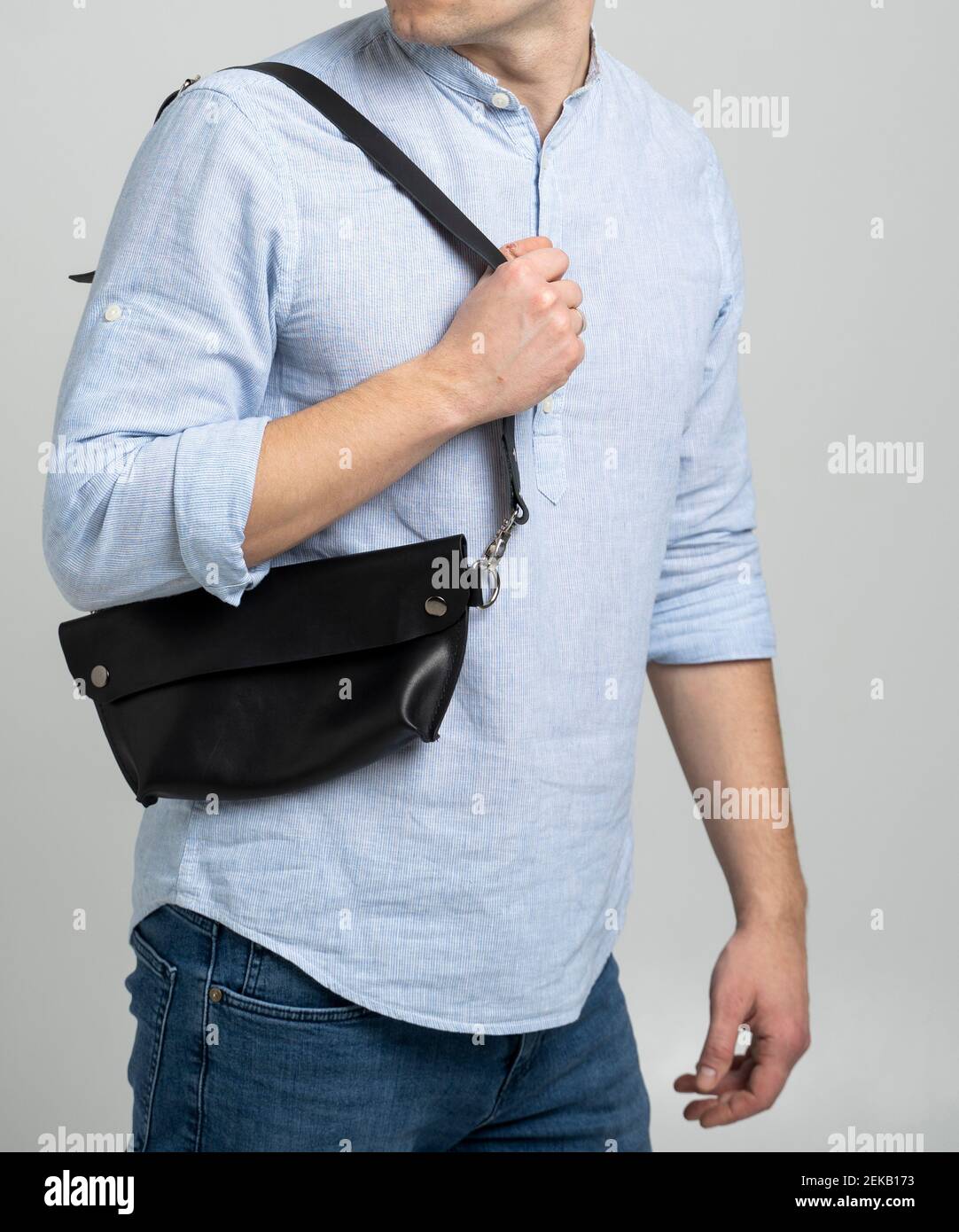 Hombre en una camisa azul con una bolsa de cuero negro hecha a mano sobre  su hombro. Bolso de bananas oscuro. Hombre en un estudio. Cómoda bolsa  pequeña para caminar Fotografía de