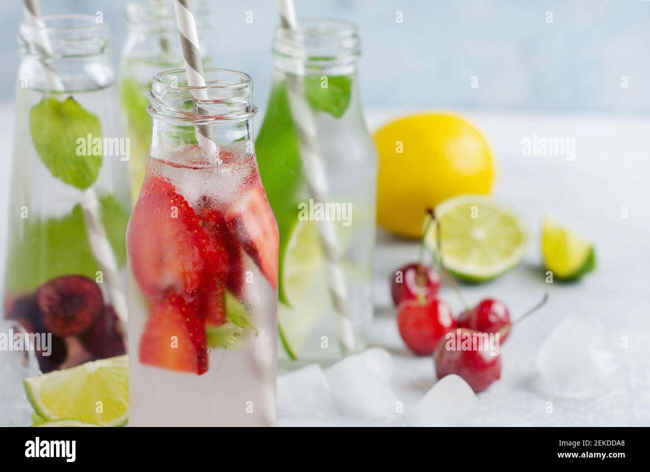 Un montón de botellas con limonada de verano refrescante con lima, fresa, cereza, pepino y hielo sobre un fondo de concreto gris Foto de stock