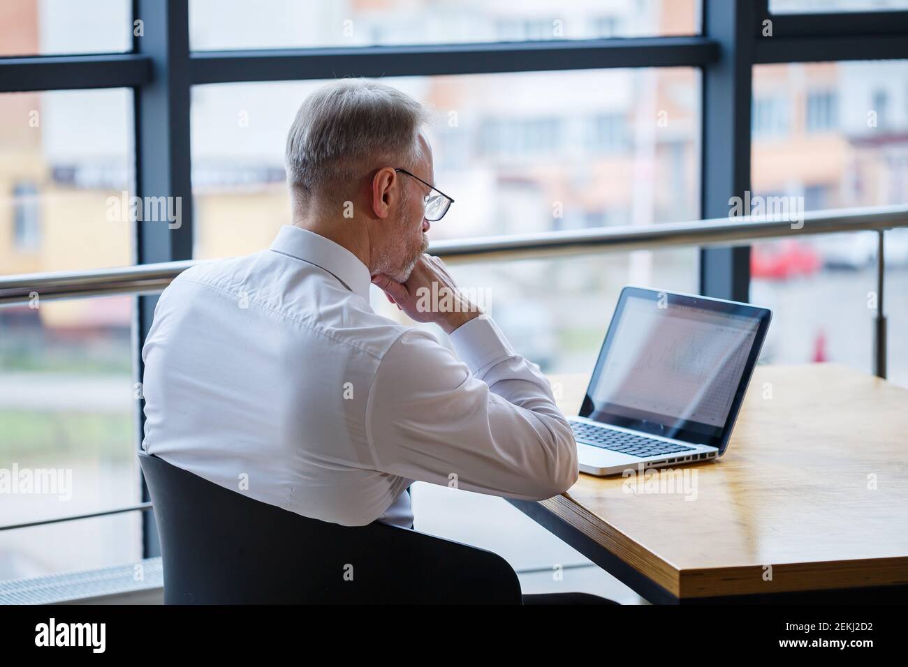 Hombre freelancer está trabajando en un café en un nuevo proyecto de negocios. Se sienta en una ventana grande en la mesa. Mira a una pantalla de portátil con una taza de café Foto de stock