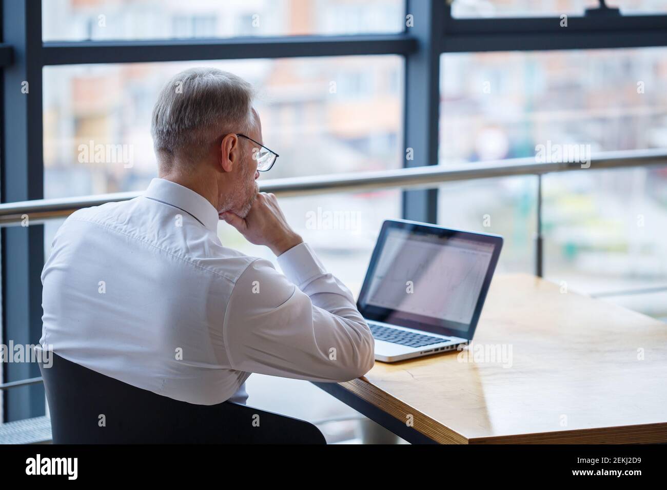 Hombre freelancer está trabajando en un café en un nuevo proyecto de negocios. Se sienta en una ventana grande en la mesa. Mira a una pantalla de portátil con una taza de café Foto de stock
