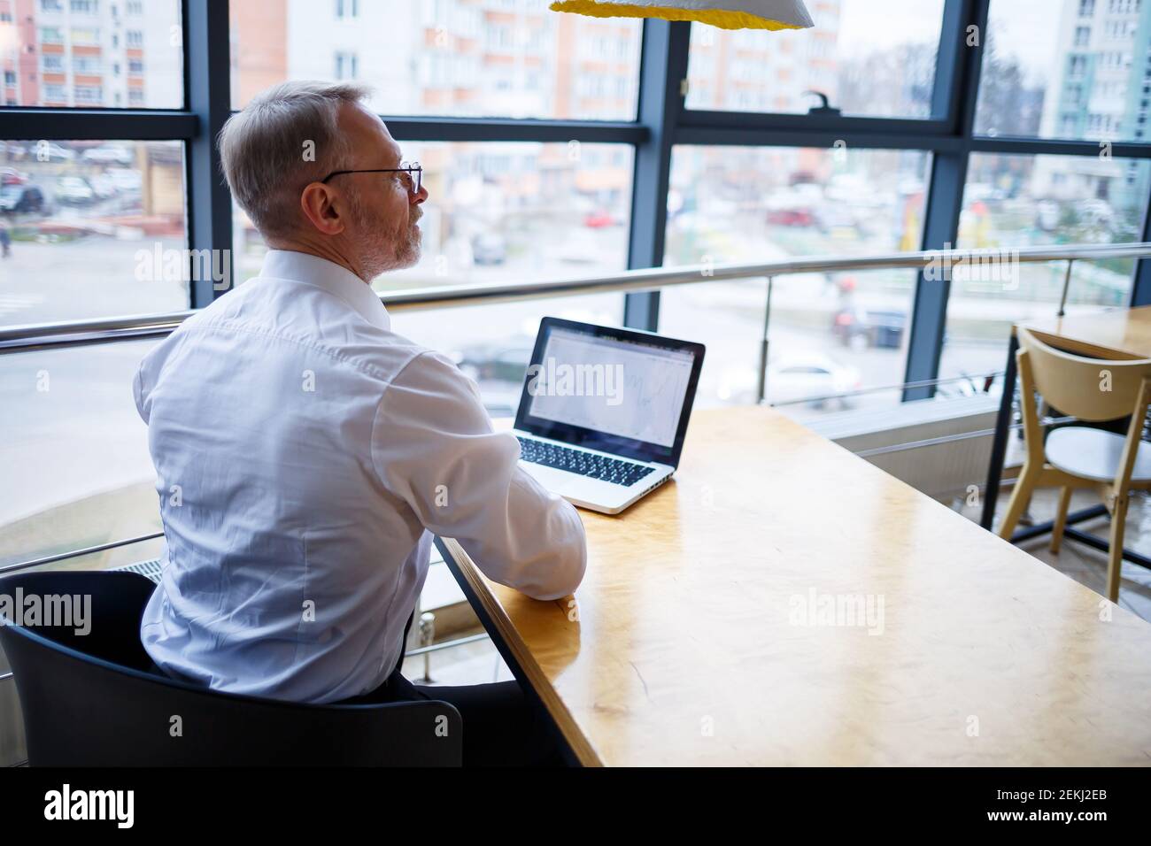 Hombre freelancer está trabajando en un café en un nuevo proyecto de negocios. Se sienta en una ventana grande en la mesa. Mira a una pantalla de portátil con una taza de café Foto de stock