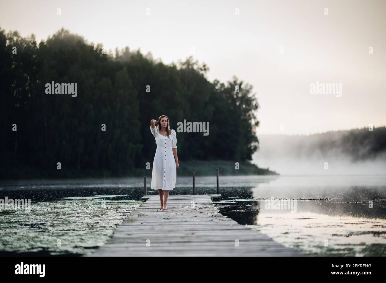 Mujer joven vestida de blanco en un lago a la mañana fría con una niebla sobre el agua. Foto de stock