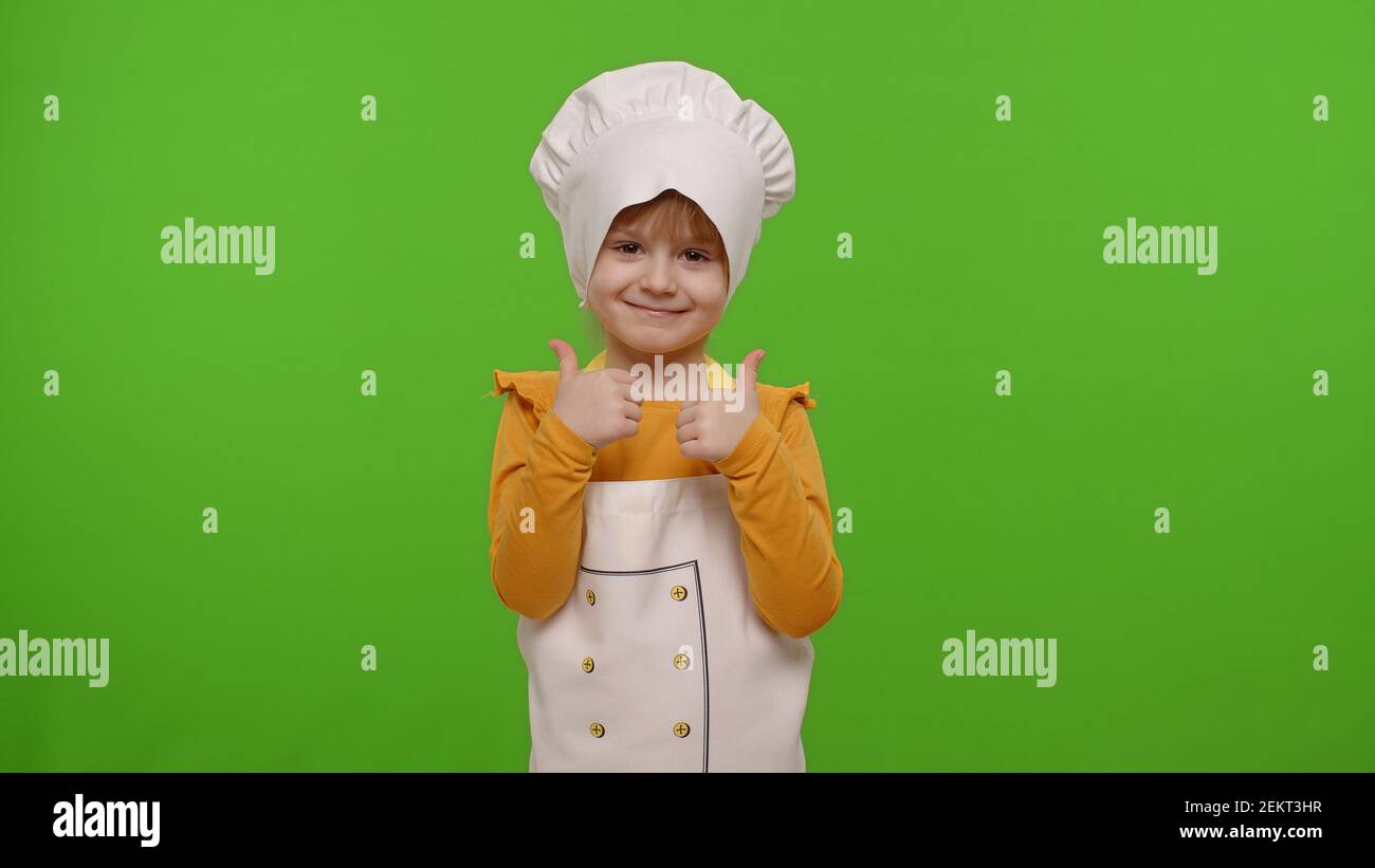 Niño niña niño vestido como cocinero profesional que muestra los pulgares hacia arriba, sonriendo, feliz a la cámara en fondo de la la croma. Nutrición, escuela de cocina,
