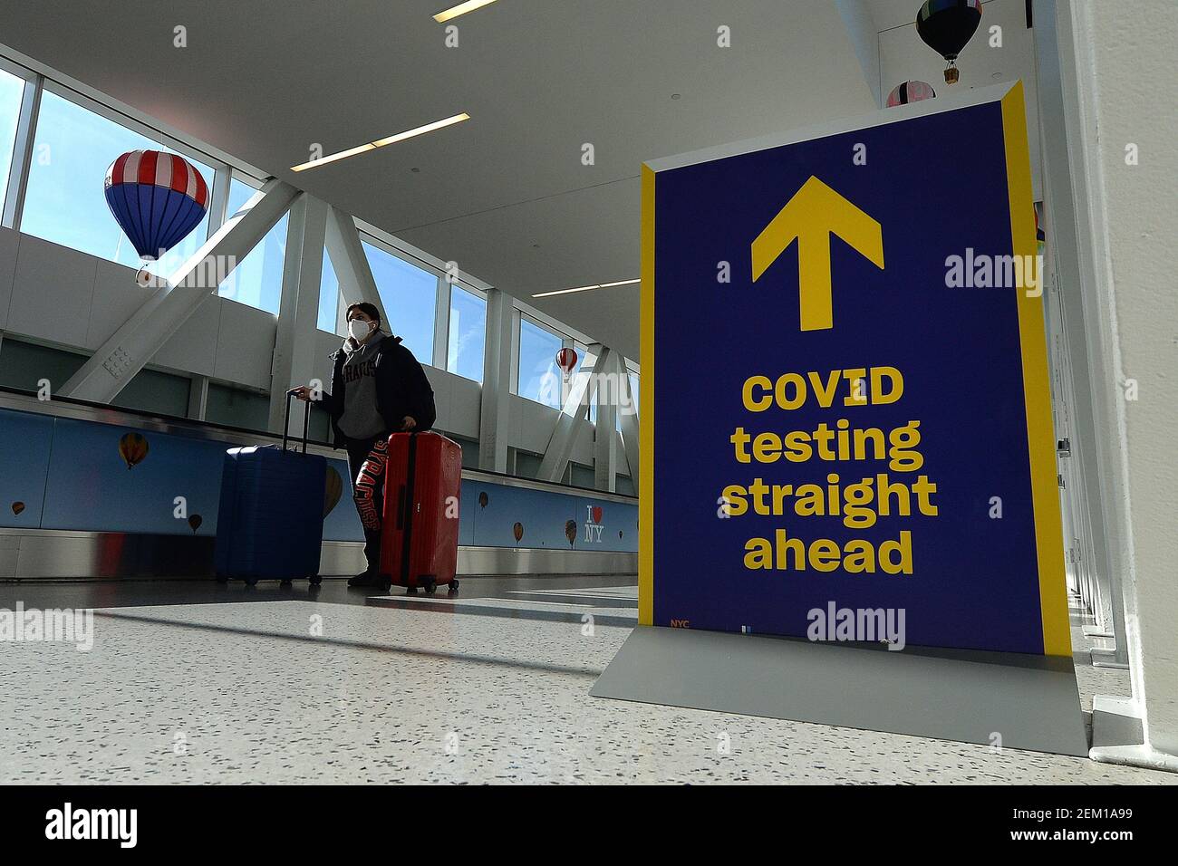 Una persona que empuja dos maletas rodantes pasa por un cartel que dirige a  la gente a un sitio de pruebas COVID-19 recientemente establecido fuera de  la Terminal 5 de Jet Blue