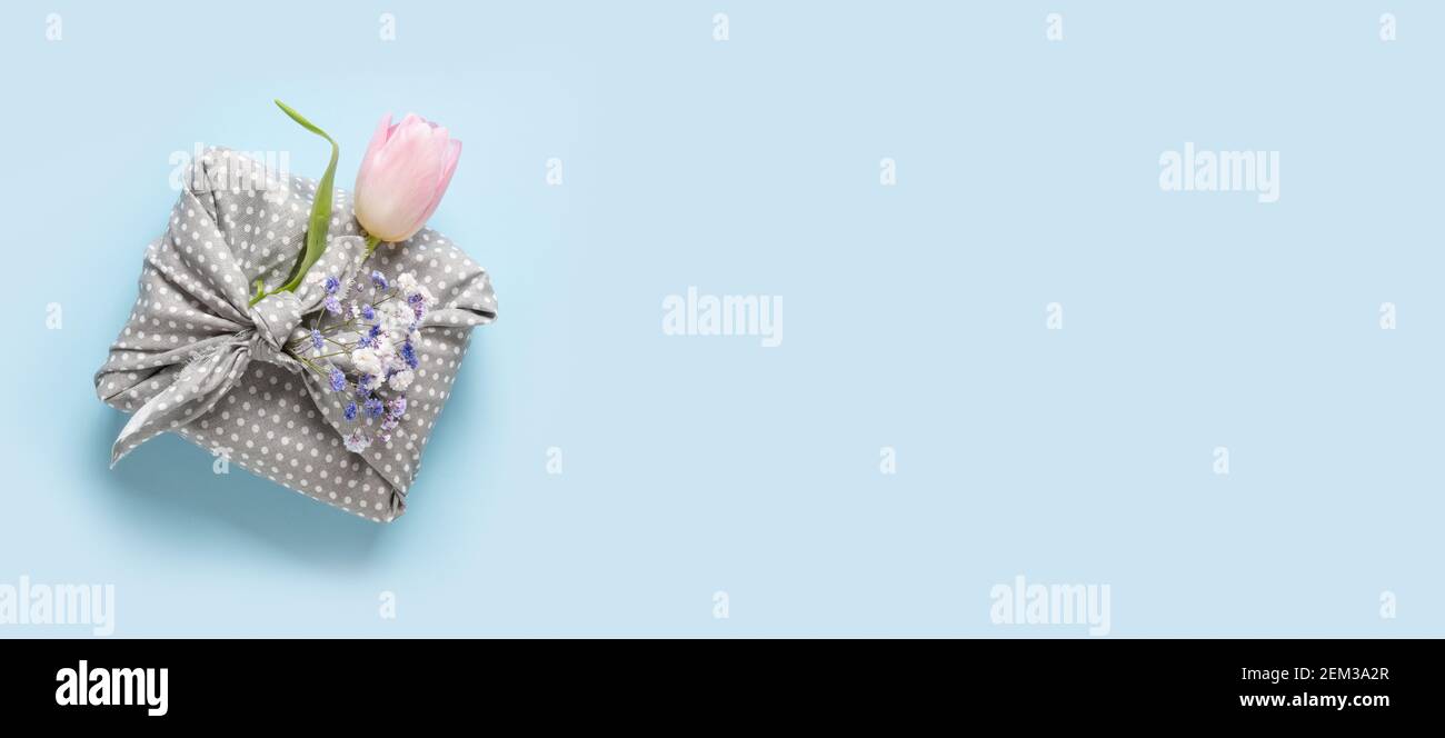 Regalo de primavera sostenible envuelto en tela gris con tulipán sobre azul. Cero residuos. Estilo tradicional japonés furoshiki. Vista desde arriba. Banner para la juerga Foto de stock