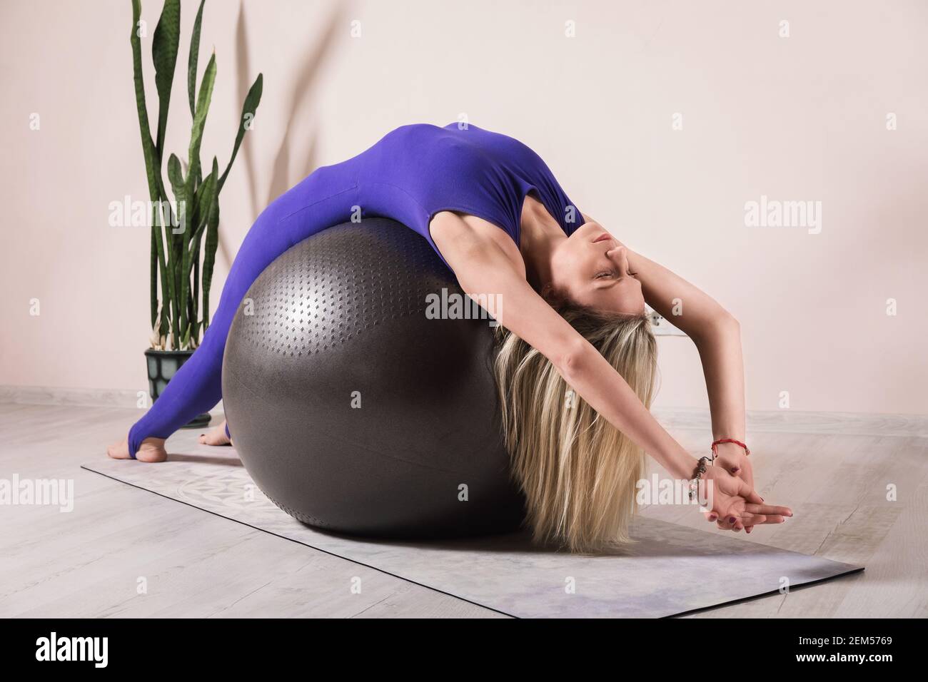 Mujer rubia en un mono deportivo cerrado de color azul practicando yoga se  sienta en un fitball con los brazos levantados en una habitación cerca de  la pared