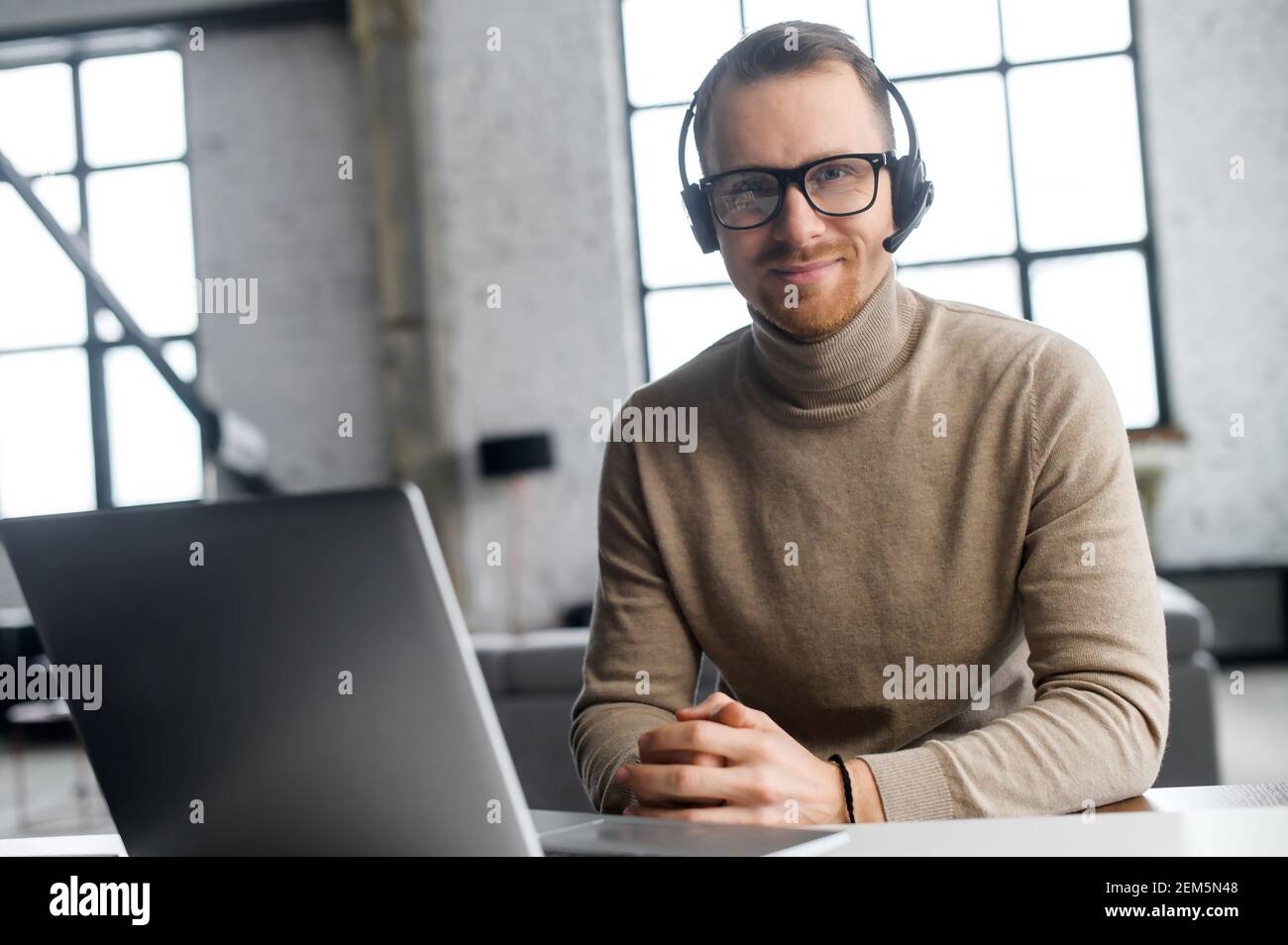 Un operador seguro con auriculares y gafas, mirando la cámara, sentado en el portátil, trabajador del año, disfruta de su trabajo, lugar de trabajo, escuchando al cliente atentamente, puede resolver su solicitud Foto de stock