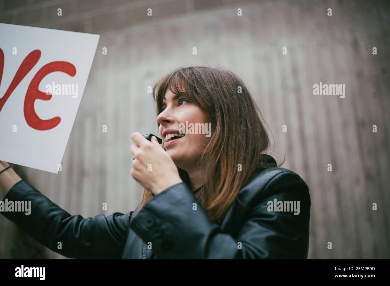 Mujer activista sonriente con silbato sosteniendo un cartel contra la pared Foto de stock