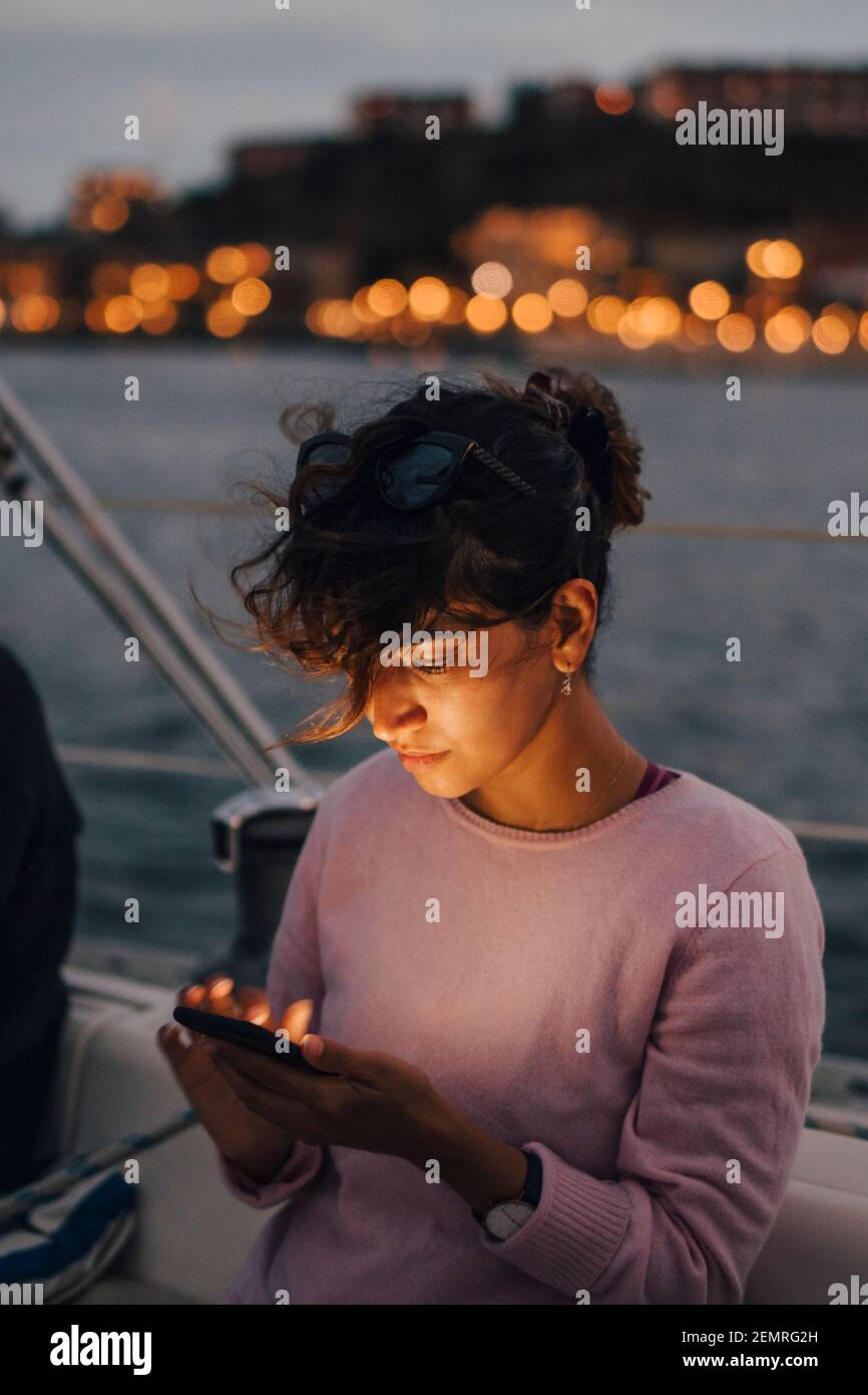 Mujer adulta mediana usando el teléfono inteligente mientras se sienta en el barco durante la puesta de sol Foto de stock