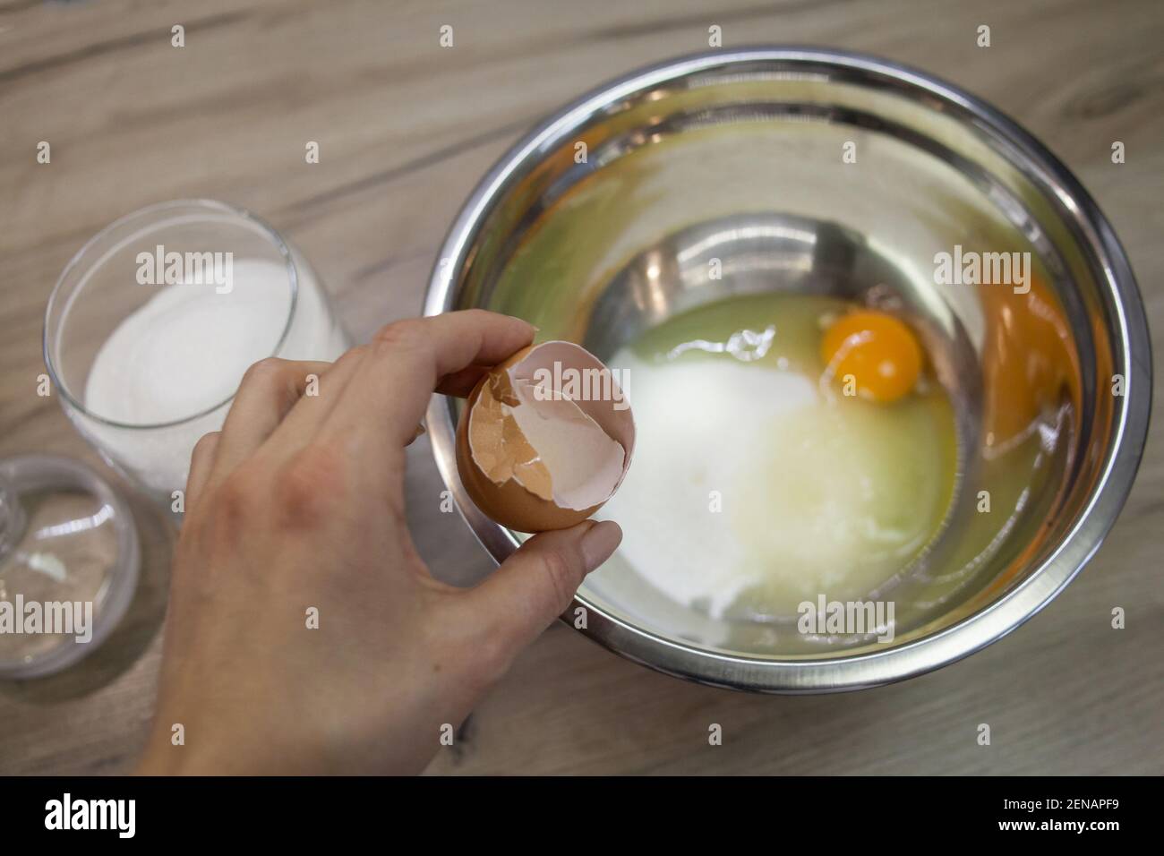 la mano sostiene la cáscara de huevo contra el fondo de un tazón metálico con azúcar y huevo. el segundo paso para hacer muffins, pastel, galletas. proceso de horneado ste Foto de stock